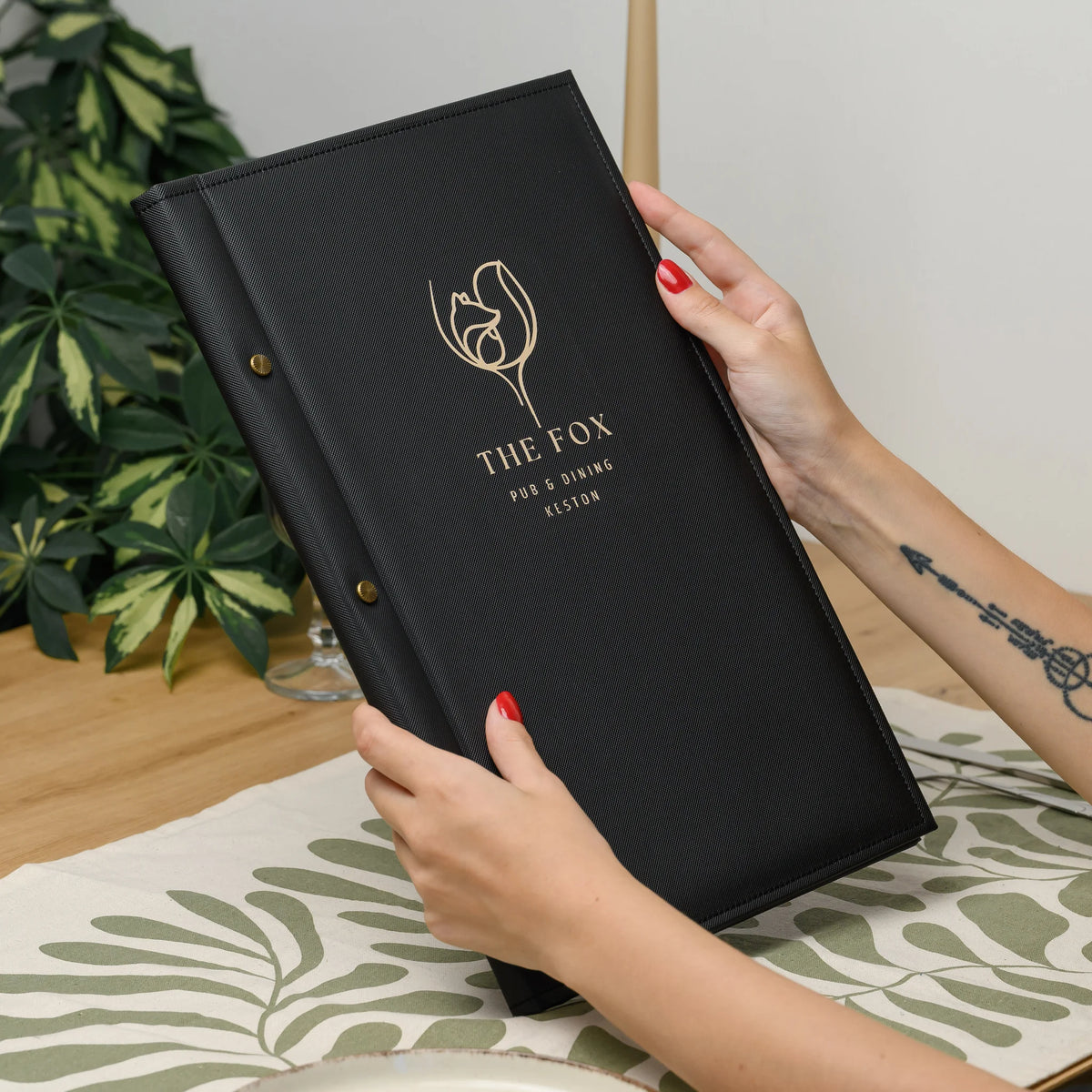 Hands holding a closed black faux leather menu cover with a gold printed logo and brass screw heads on the spine