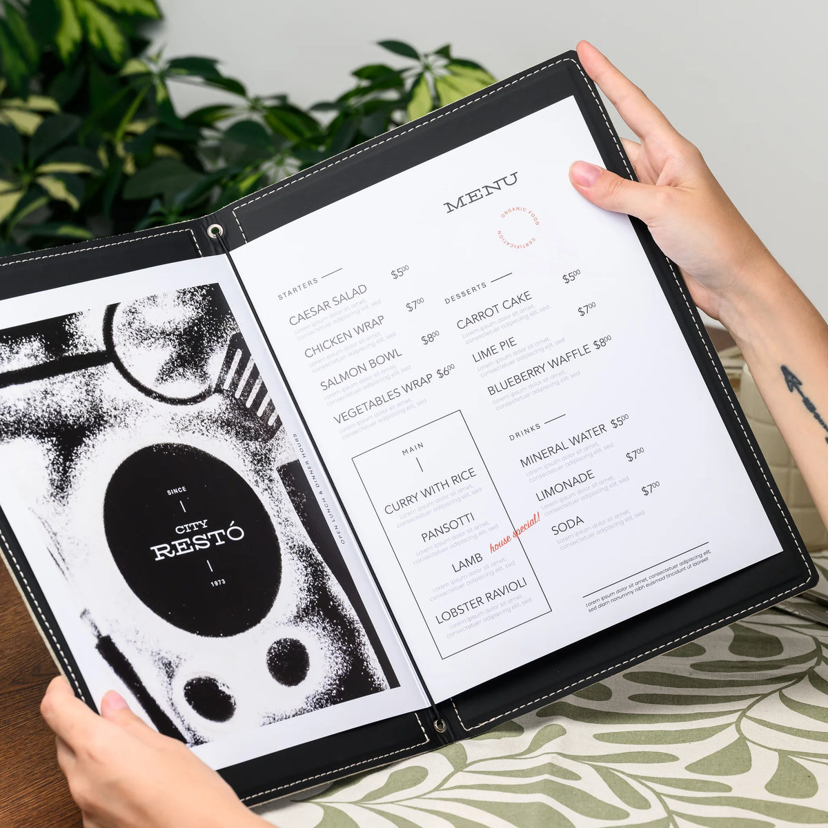 Hands holding an open menu folder showing printed menu pages secured by an elastic cord, green plants behind