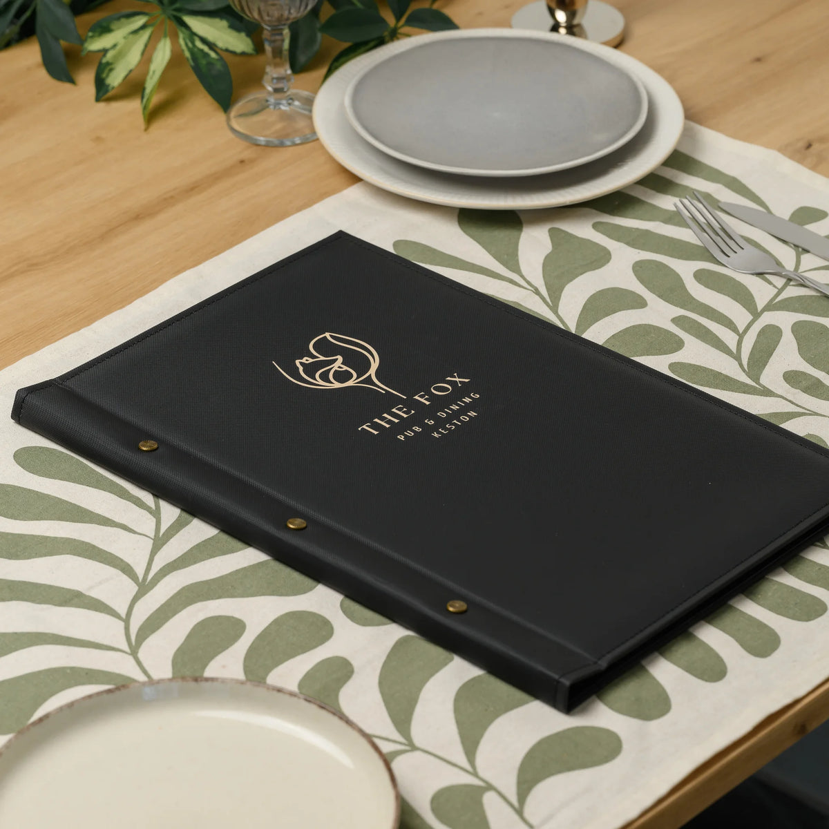 Closed black textured menu cover with a gold printed logo and two decorative brass screws lying on a table runner