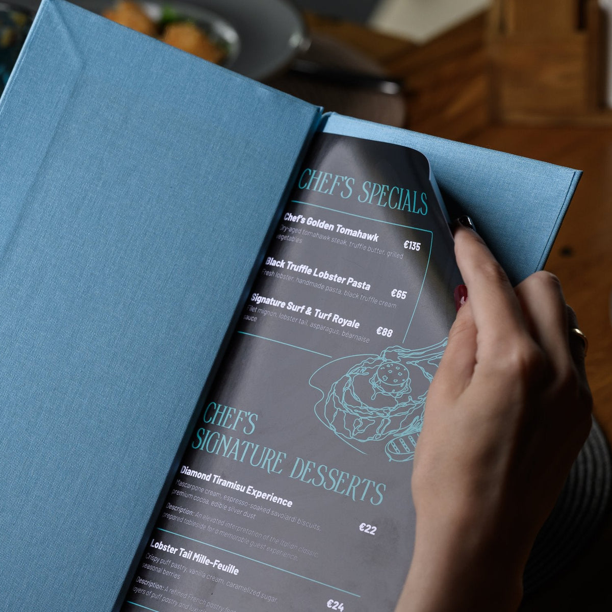 Hand opens a light-blue textile menu folder revealing a dark inner page with white script text on wood