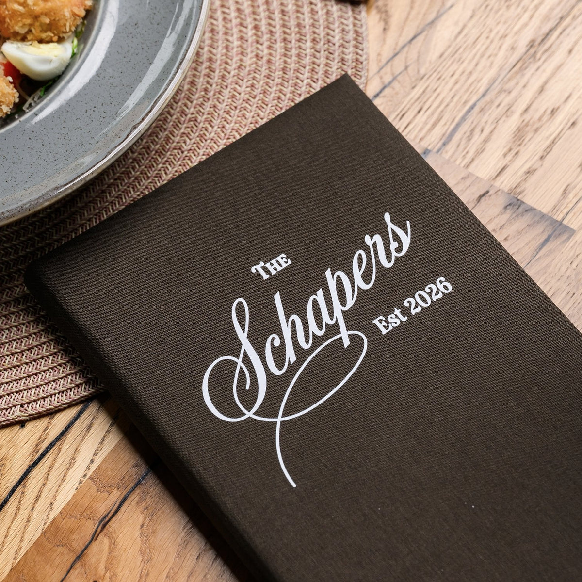 Closed espresso-brown textile menu folder standing upright with a cursive logo beside a blurred salad plate