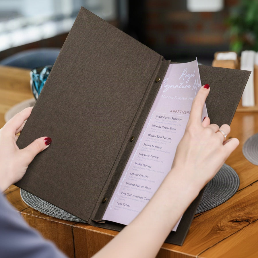Hand holds a wide open espresso-brown textile menu folder with a lavender page beside a salad plate edge