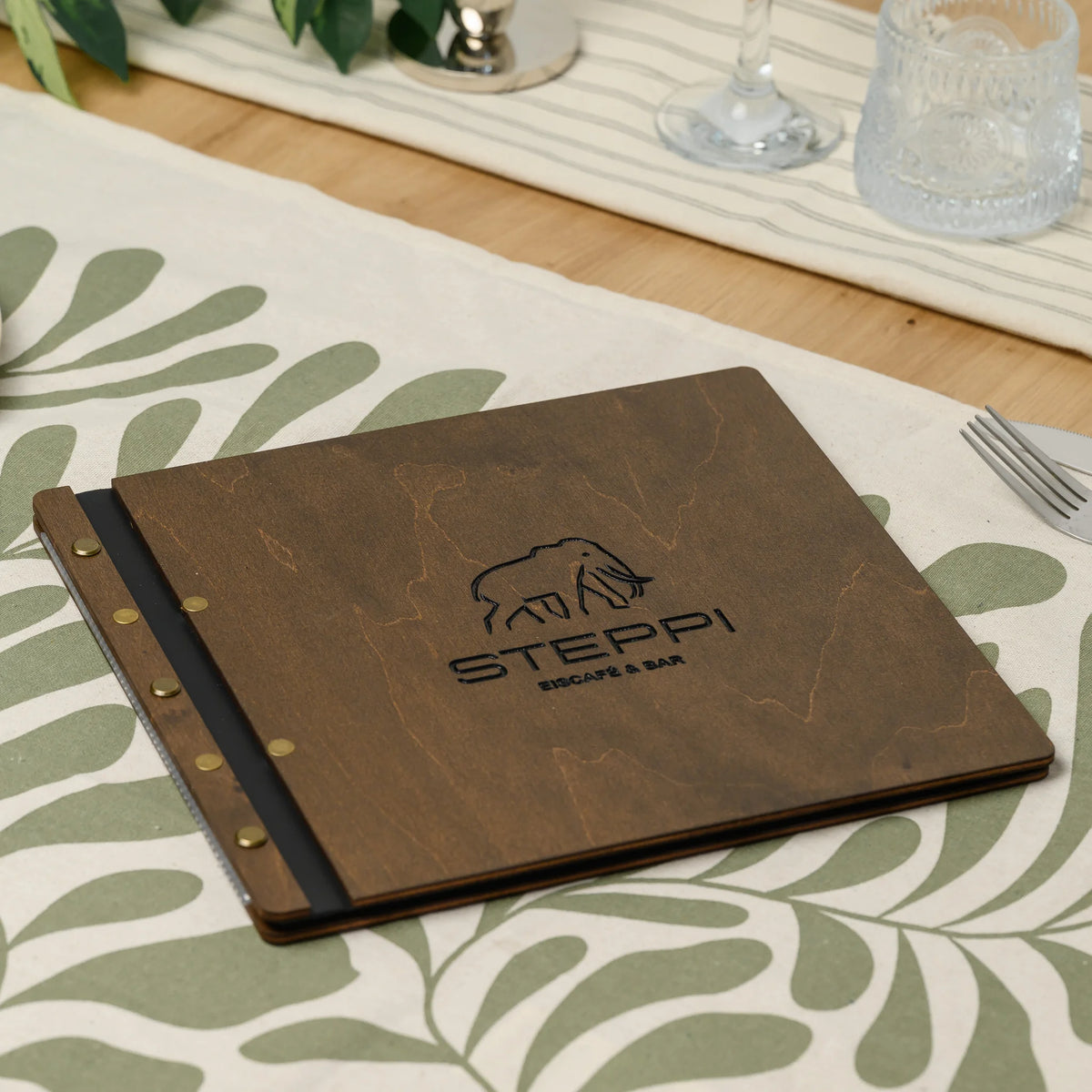 Square wooden menu cover in walnut plywood with an engraved logo and brass screws on the spine on a table runner