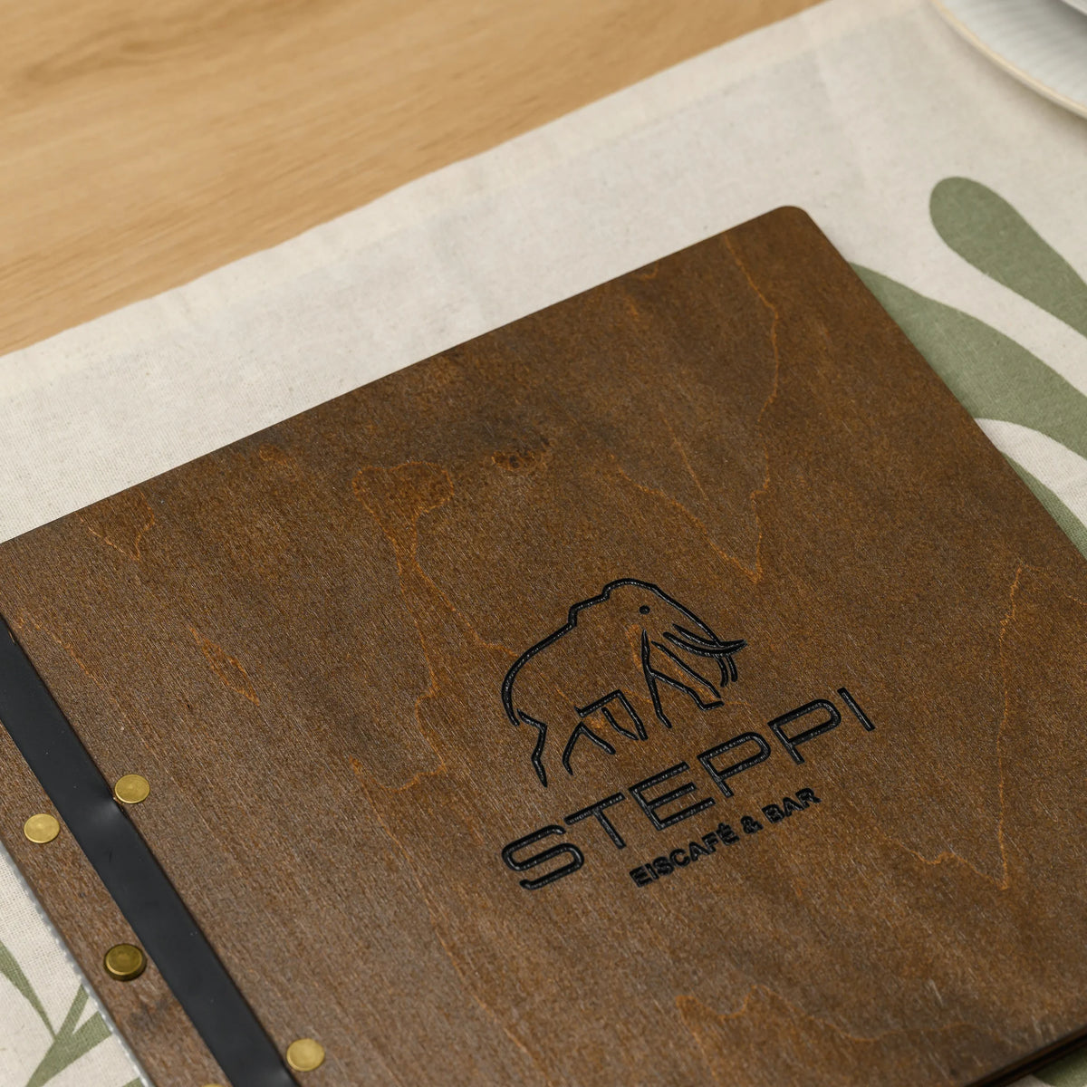 Walnut plywood menu cover with an engraved logo, black leather strap and brass screws lying on a set restaurant table