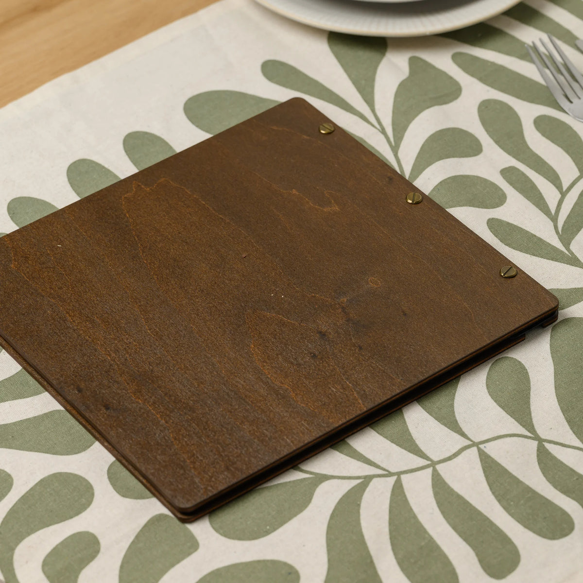 Square wooden menu cover in walnut plywood with an engraved logo and brass screws on the spine on a table runner