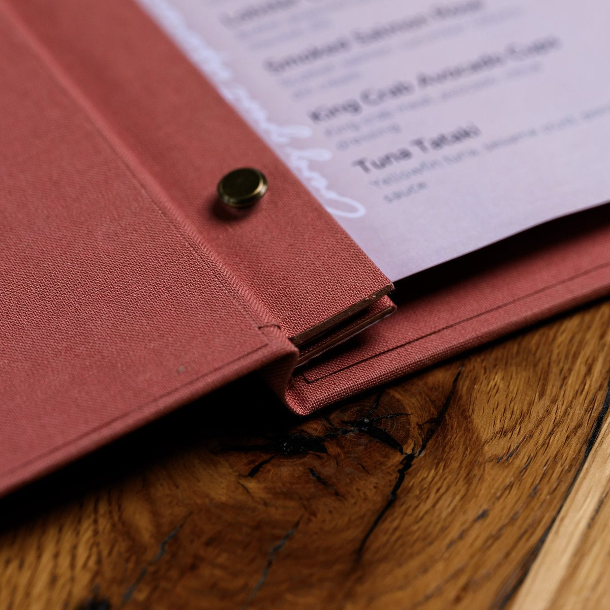 Open coral-pink textile menu folder shown from the side with a lavender page and a rivet fastener on wood