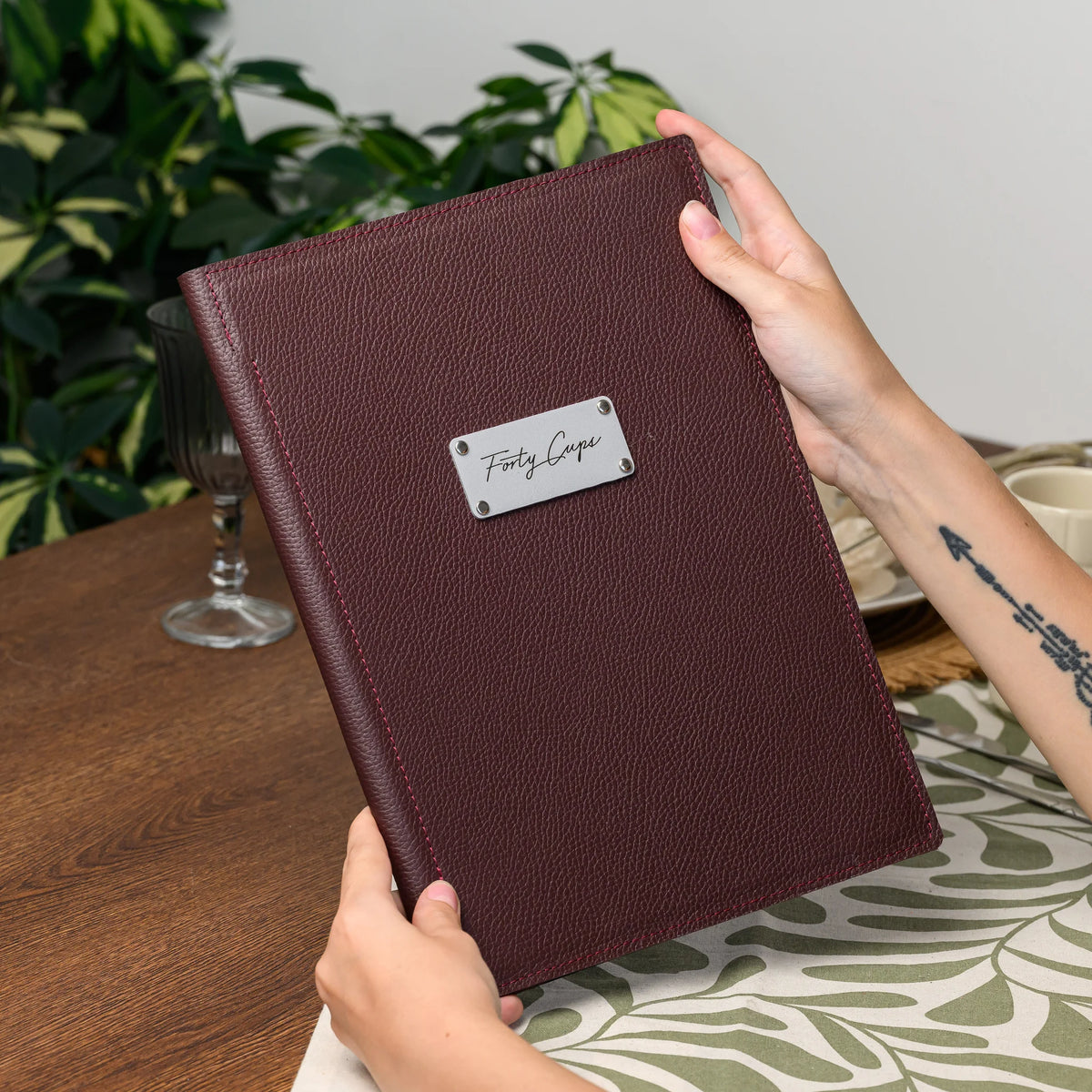 Hands holding a closed bordeaux leather menu cover with a rectangular silver logo plate in front of green plants