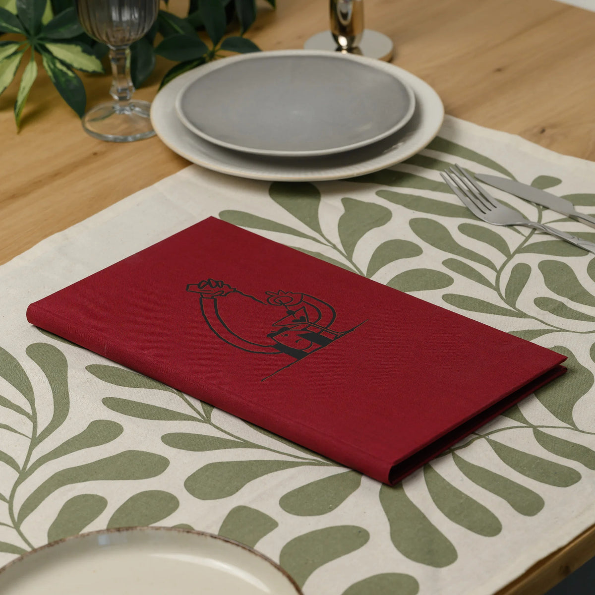 Closed red fabric menu cover with a black DTF printed line illustration lying on a patterned runner near plates