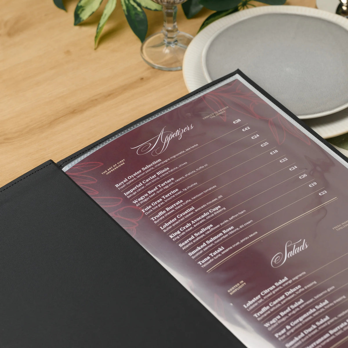Open black faux leather menu cover on a restaurant table next to plates, with a dark printed menu in a clear sleeve