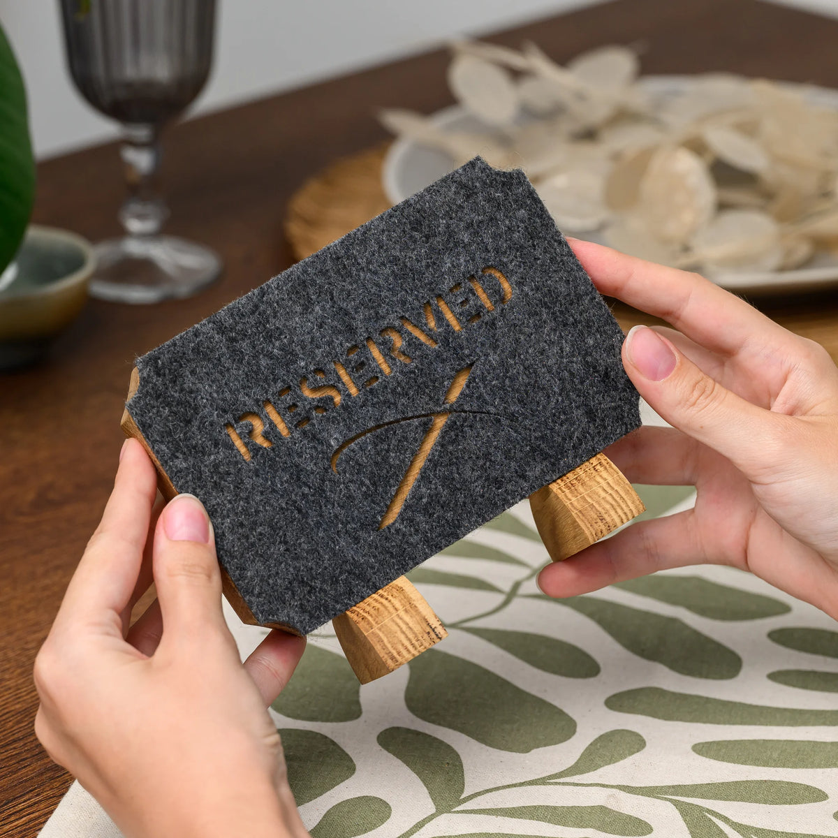 A hand holding a Reserved table sign, showing its felt cutout lettering and oak leg stand, viewed up close