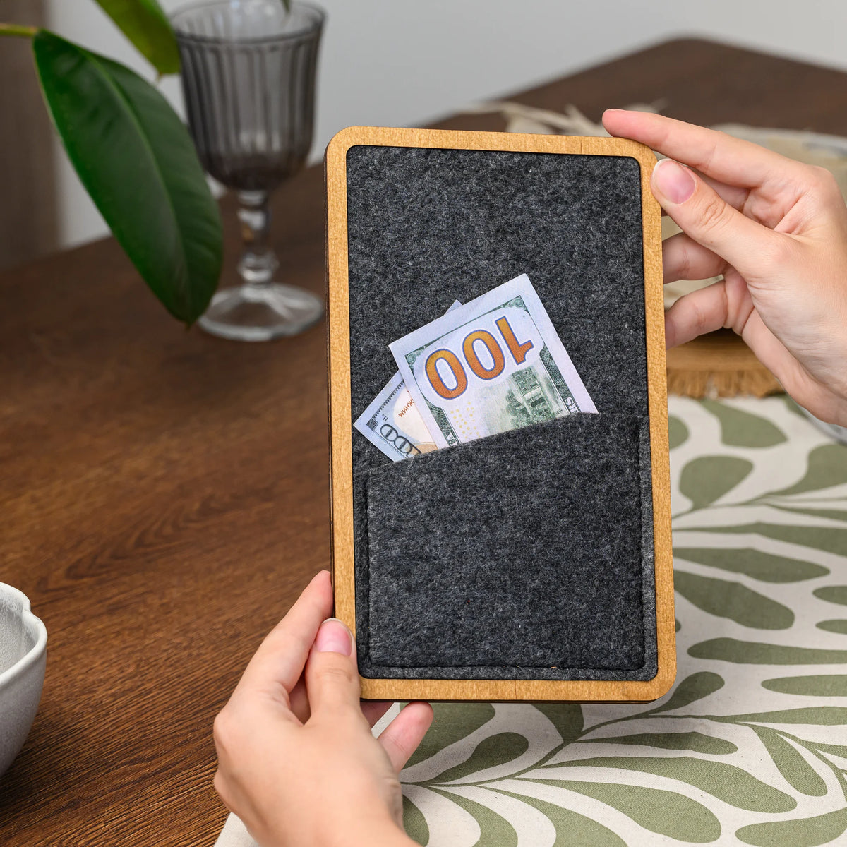 Hands holding a wood-framed bill presenter with a banknote tucked into its stitched felt pocket closely 
