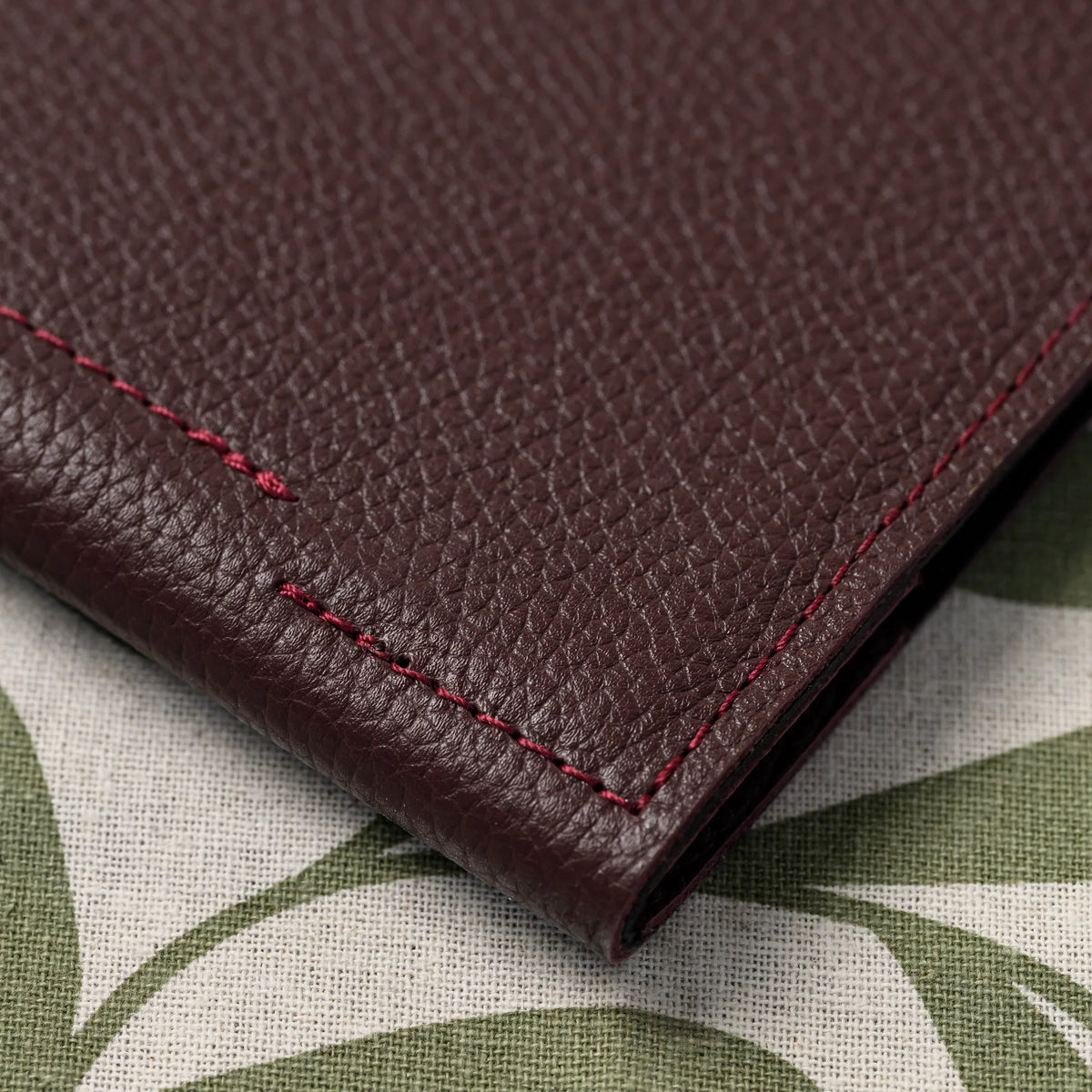 Macro of bordeaux pebbled leather cover corner with contrasting red stitching lying on leaf-print linen cloth
