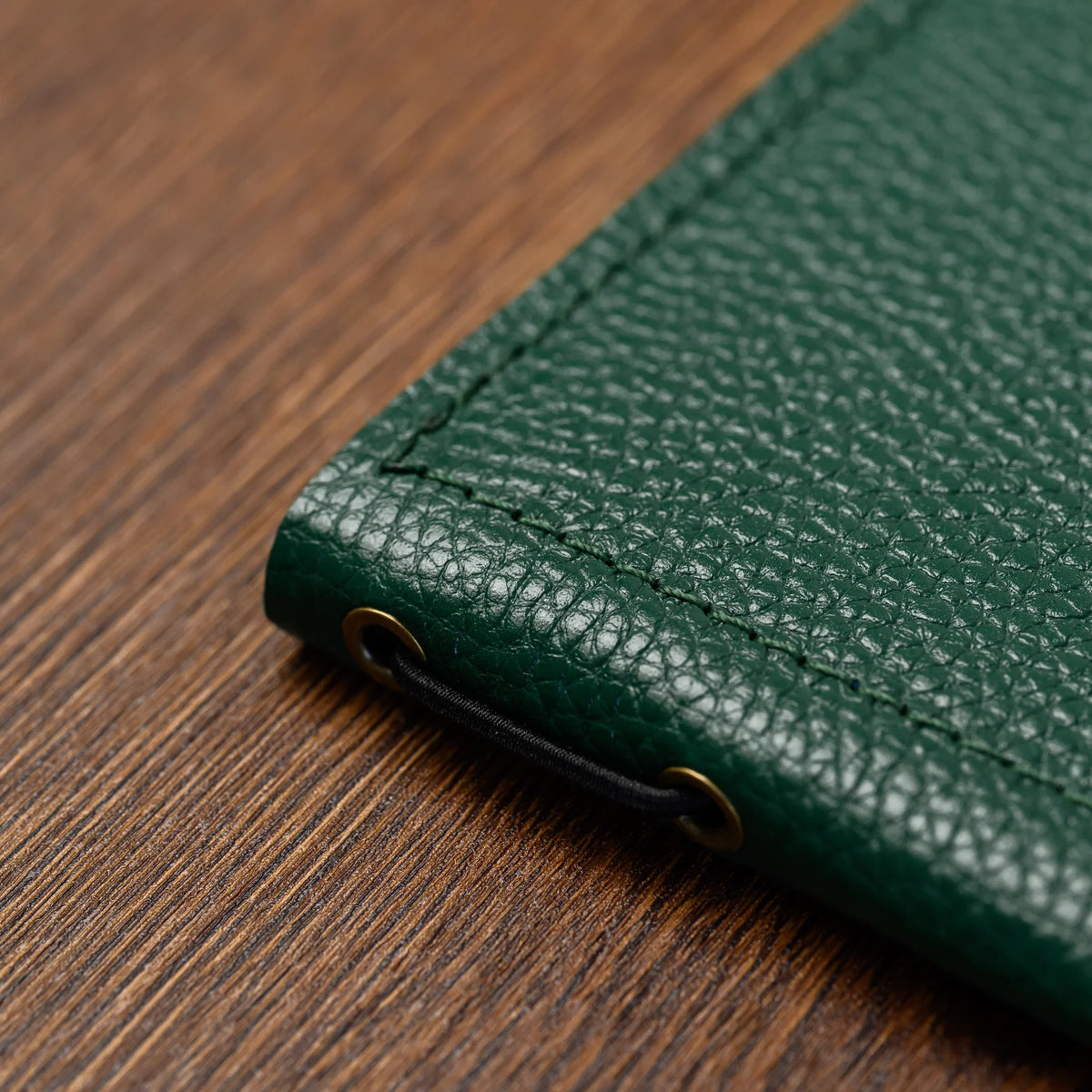 Close-up of green pebble-grain leather folder spine with two brass eyelets and black elastic cord on wood table