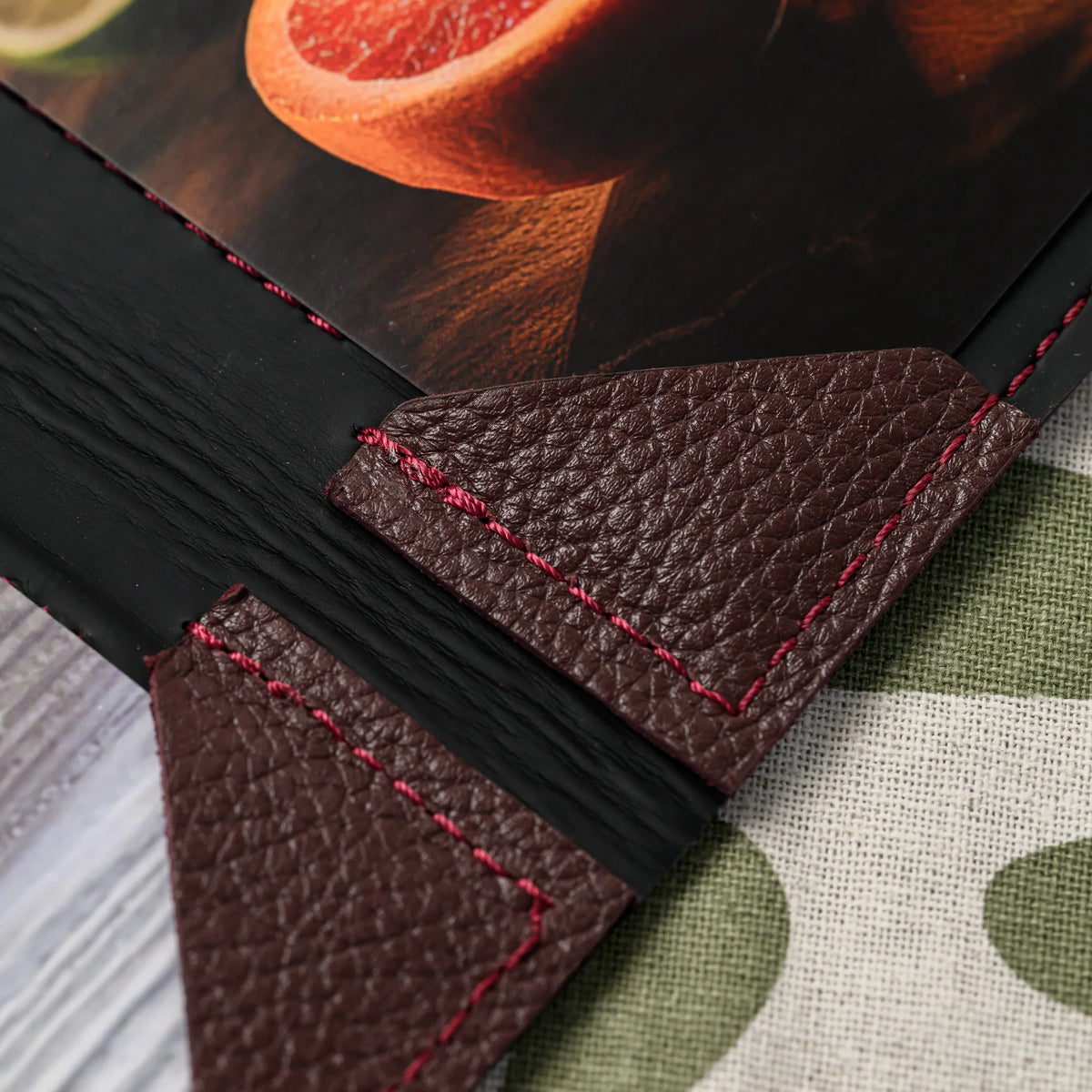 Macro of two bordeaux leather corner mountings with red stitching beside the black spine and a photo menu sheet