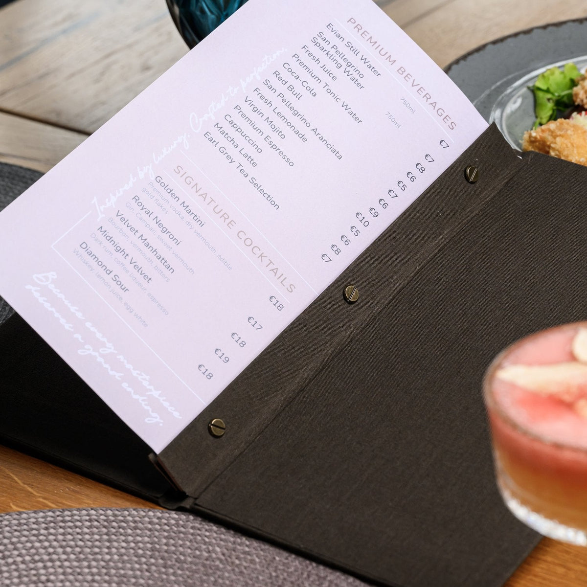 Wide open espresso-brown textile menu folder with a lavender menu page beside a salad plate and cocktail