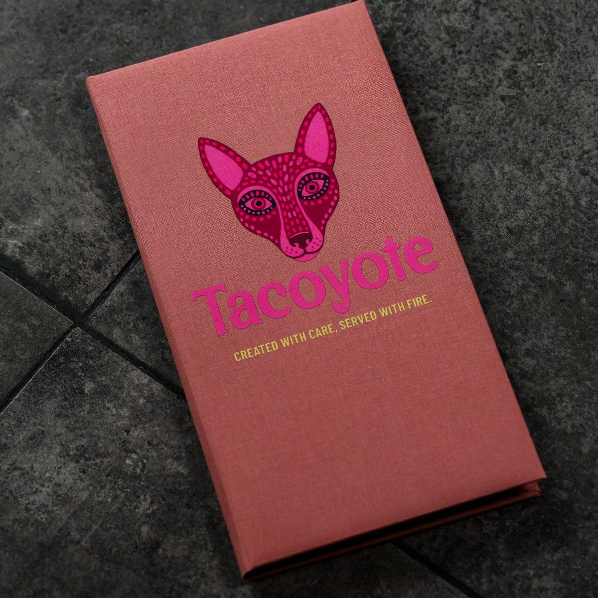 Closed coral-pink textile menu folder standing upright with a fox graphic on a dark grey stone tile floor