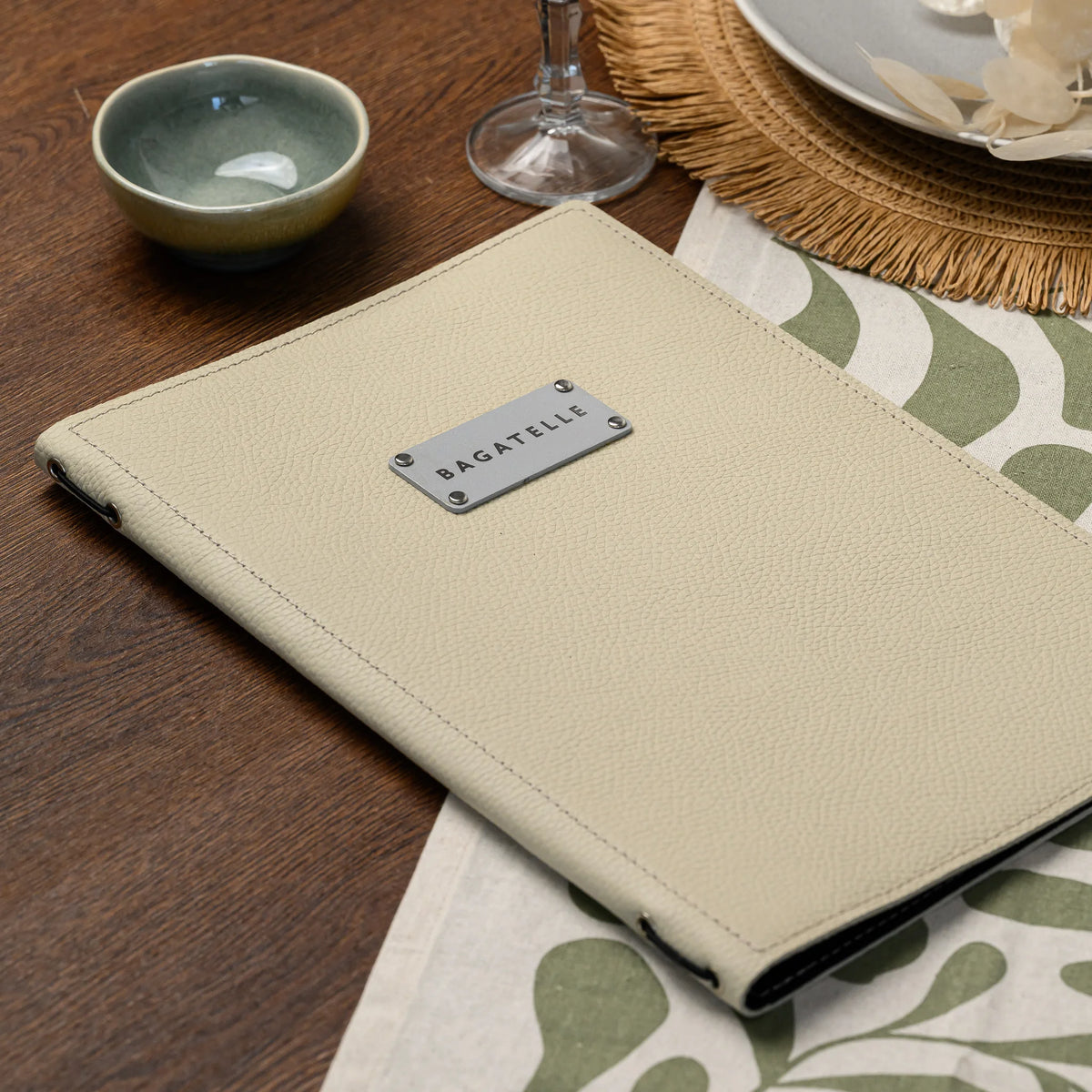 Closed beige leather menu folder with rectangular silver logo plate on a set table beside a bowl and wine glass