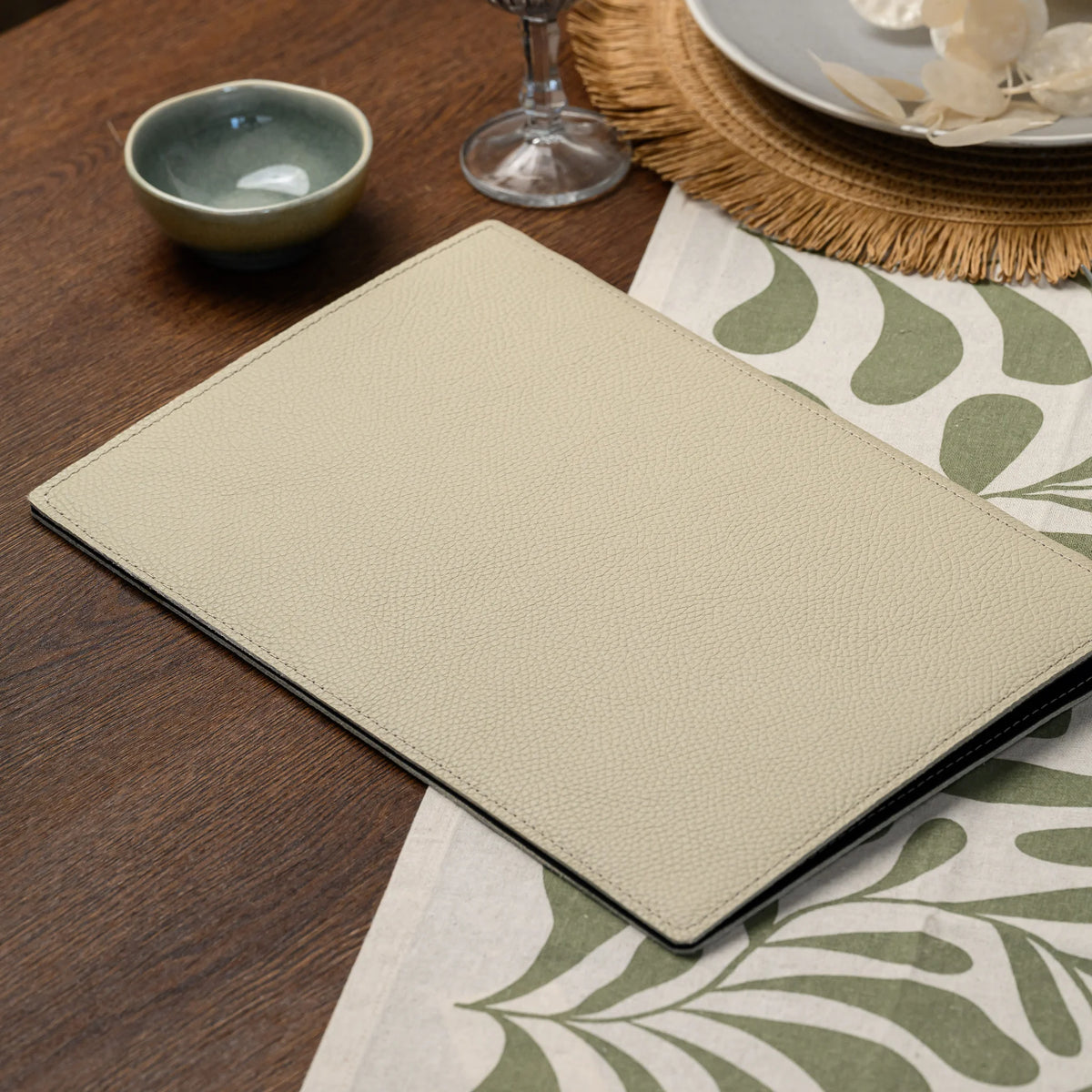 Plain beige leather menu folder lying flat on a leaf-print table runner next to a place setting and glassware