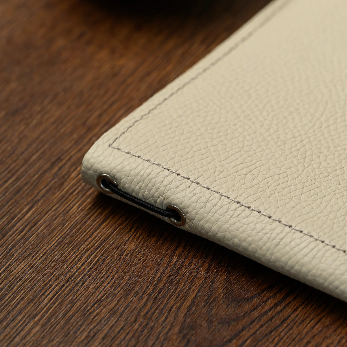 Macro of beige milled leather spine edge with two silver eyelets and black elastic cord on a dark wood table