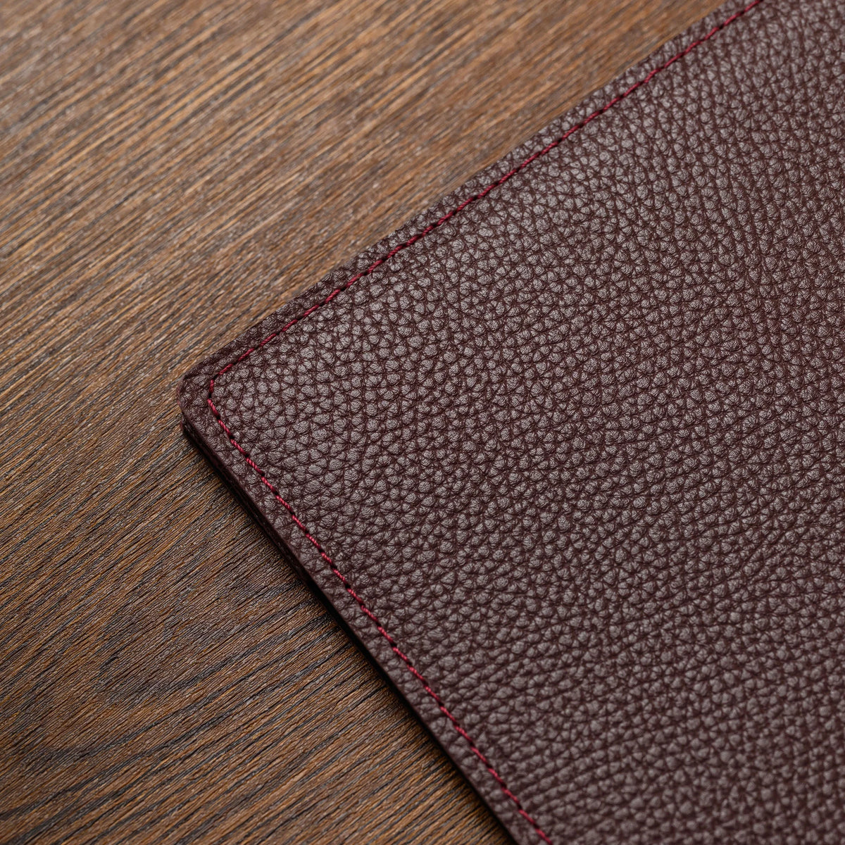 Close-up of bordeaux milled leather cover corner showing fine pebble grain and red edge stitching on wood