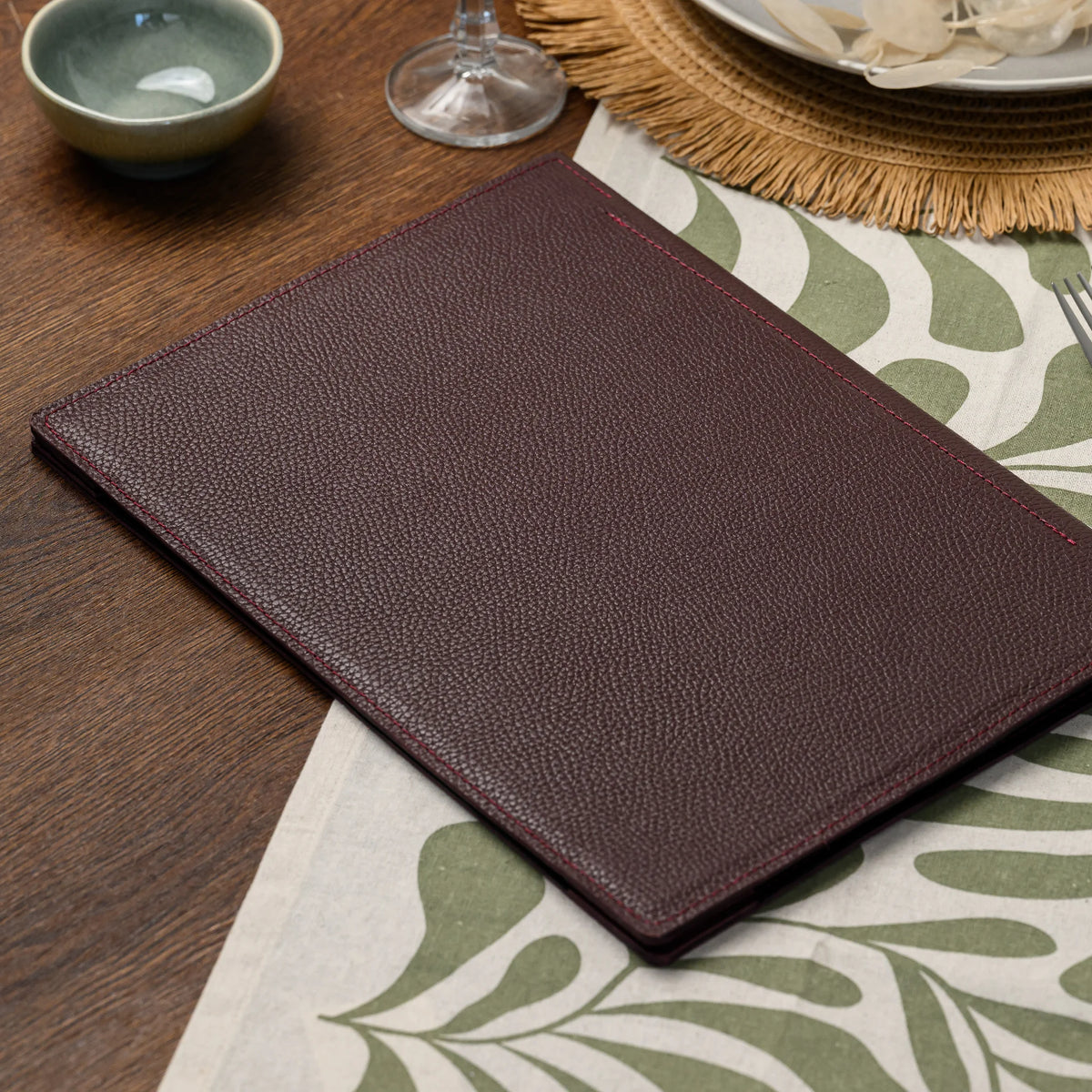 Plain bordeaux leather menu cover without a logo plate on a leaf-print table runner near a place setting
