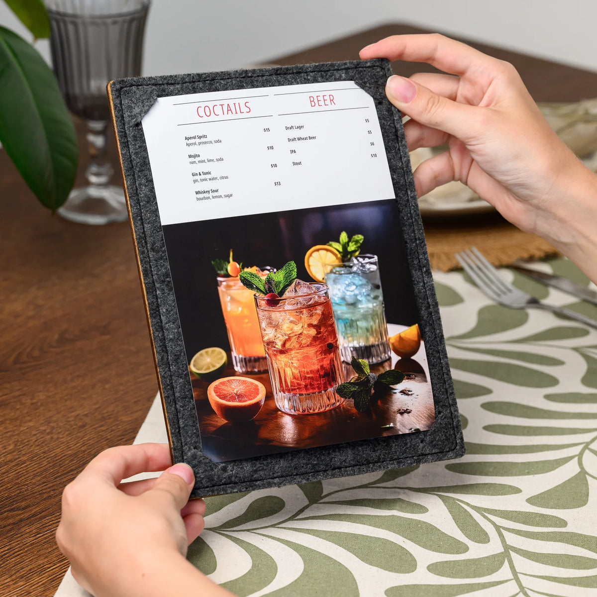 Hands holding a felt menu clipboard at an angle, showing the printed page held by its stitched corner slot 