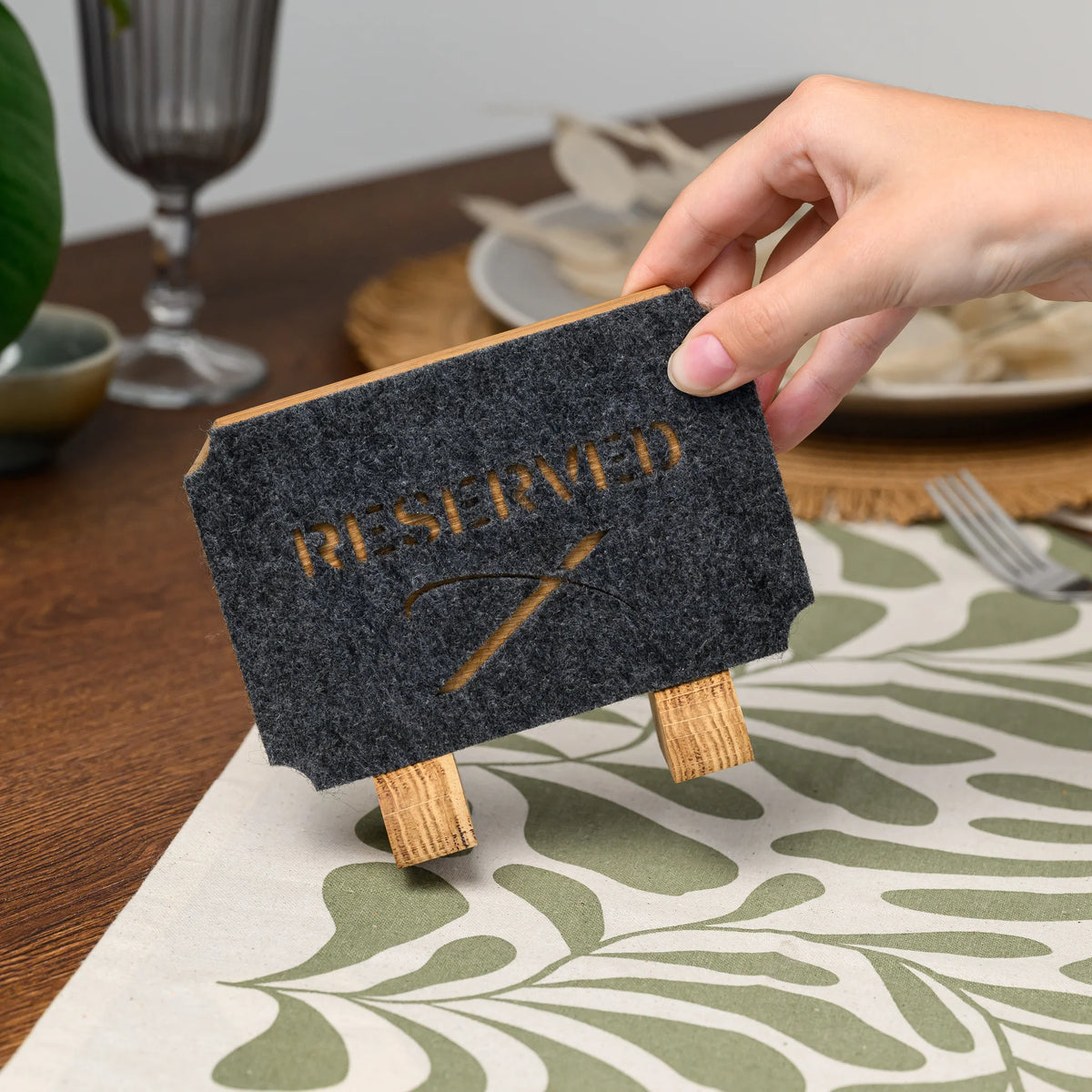 A hand holding a Reserved table sign at an angle, showing the felt lettering and its oak stand legs closely 