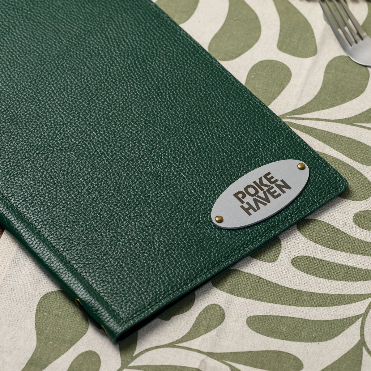 Green leather menu folder on a patterned table runner with an oval silver plate fixed by two brass rivets