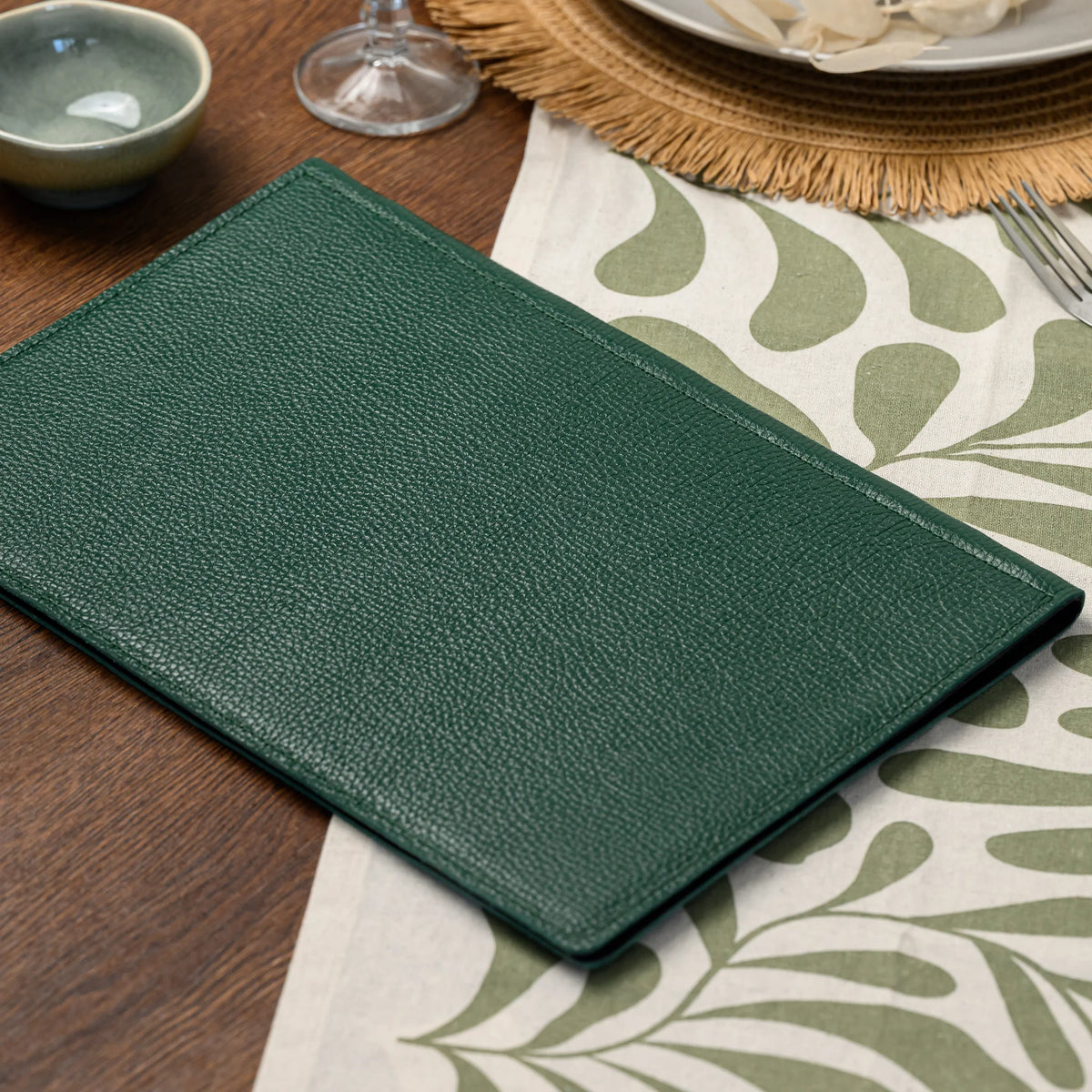 Plain green leather menu cover without a logo plate lying on a leaf-print table runner next to a ceramic bowl