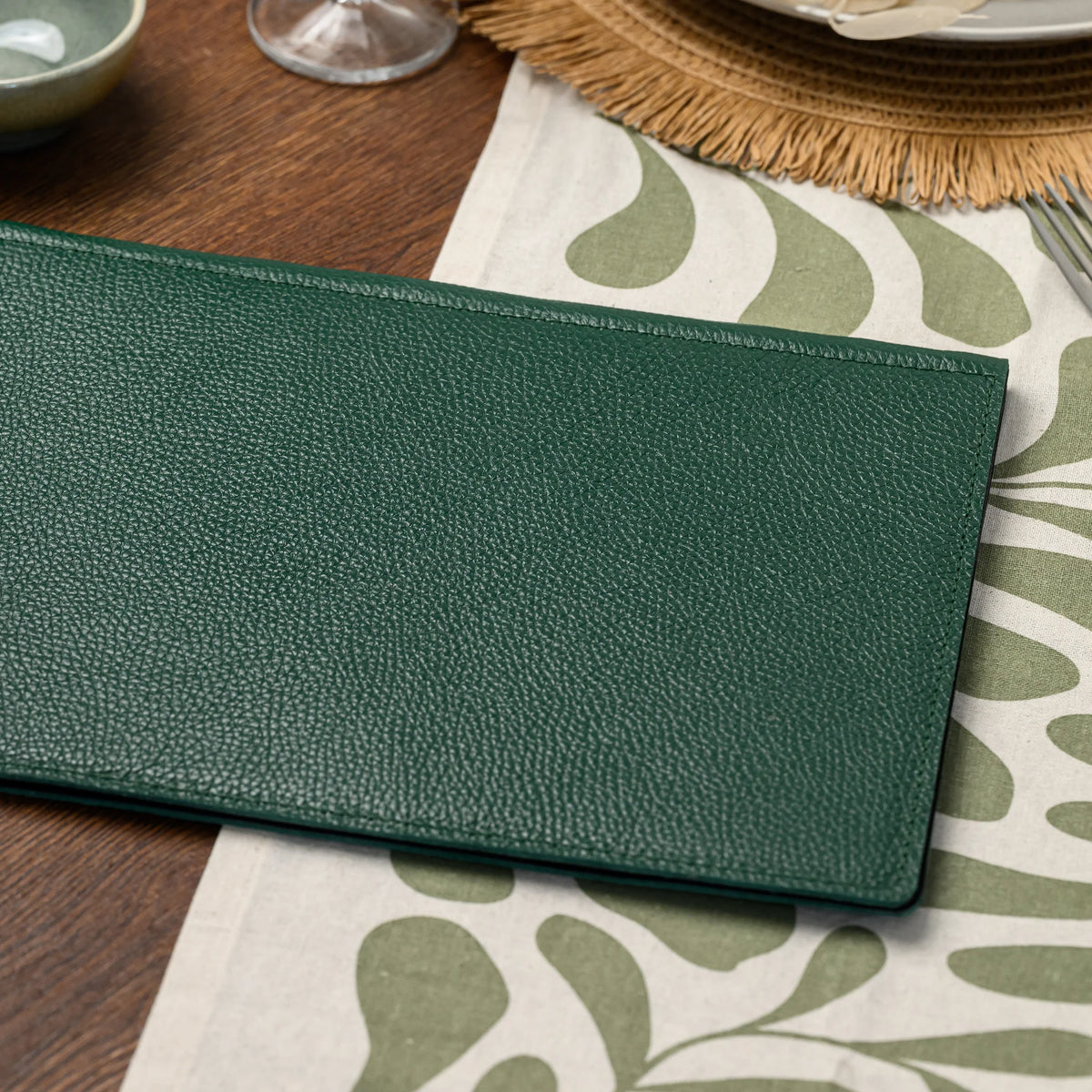 Wide crop of plain green milled leather menu cover lying flat on a table runner, stitched border visible