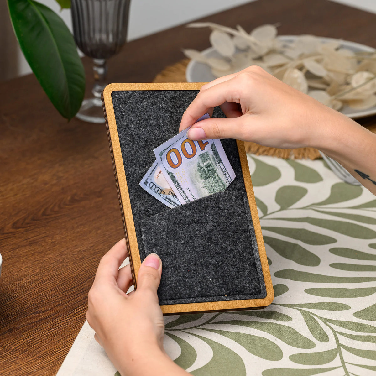 Hands placing a folded banknote into the felt pocket of a wood-framed bill presenter, viewed closely 