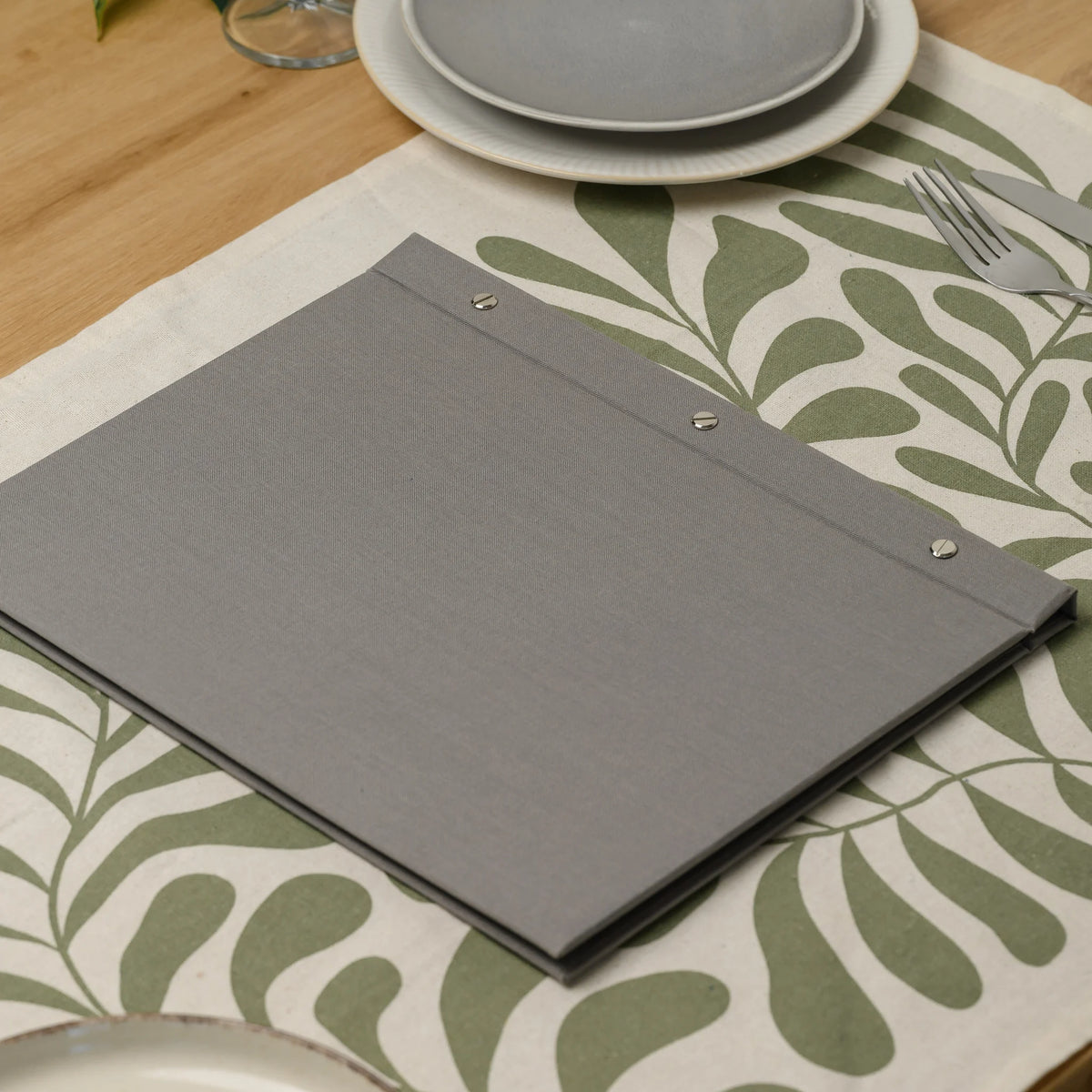 Closed gray fabric menu cover with three chrome screws along the spine lying on a set table next to plates