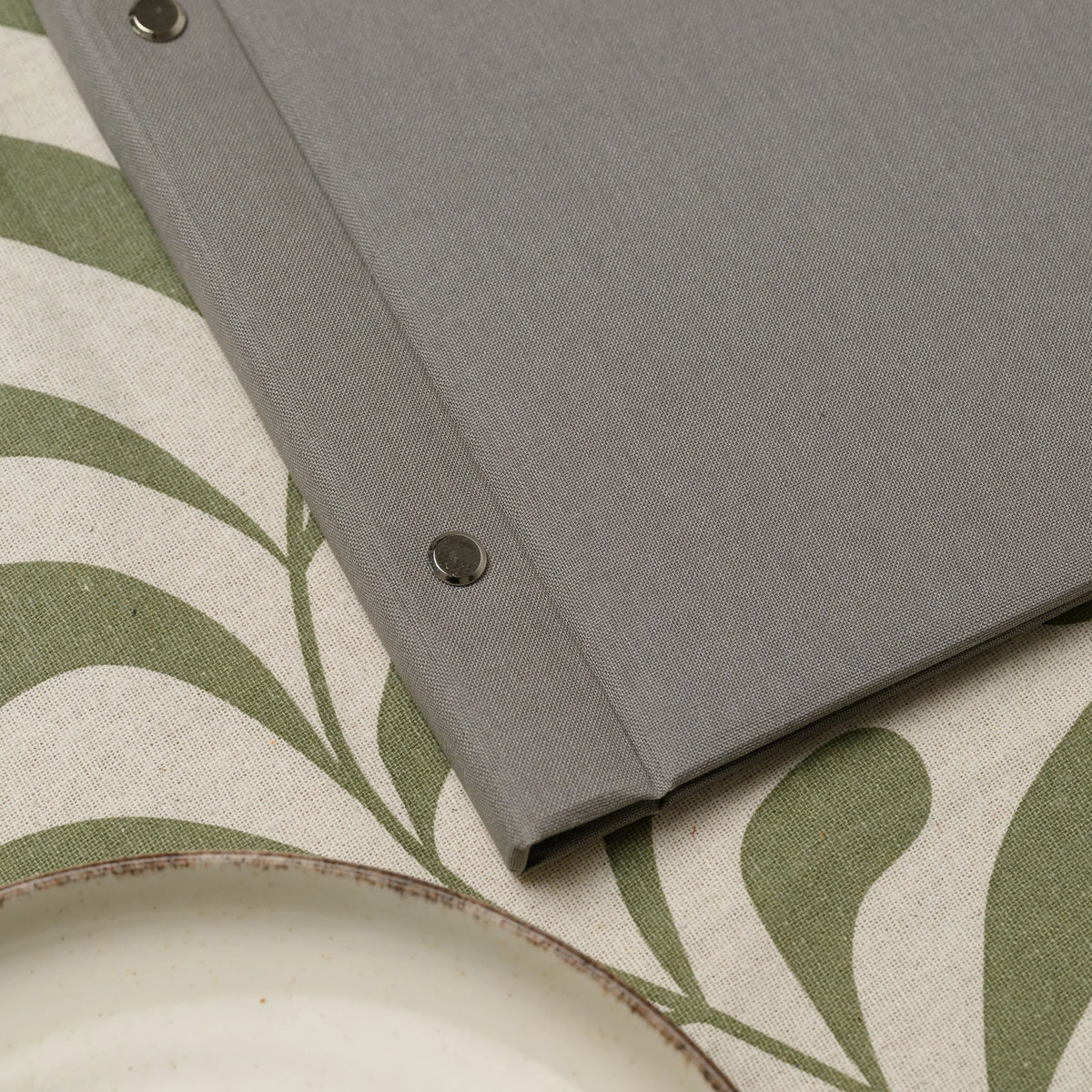 Close-up of a chrome screw head on the spine of a gray fabric menu cover next to the rim of a ceramic plate