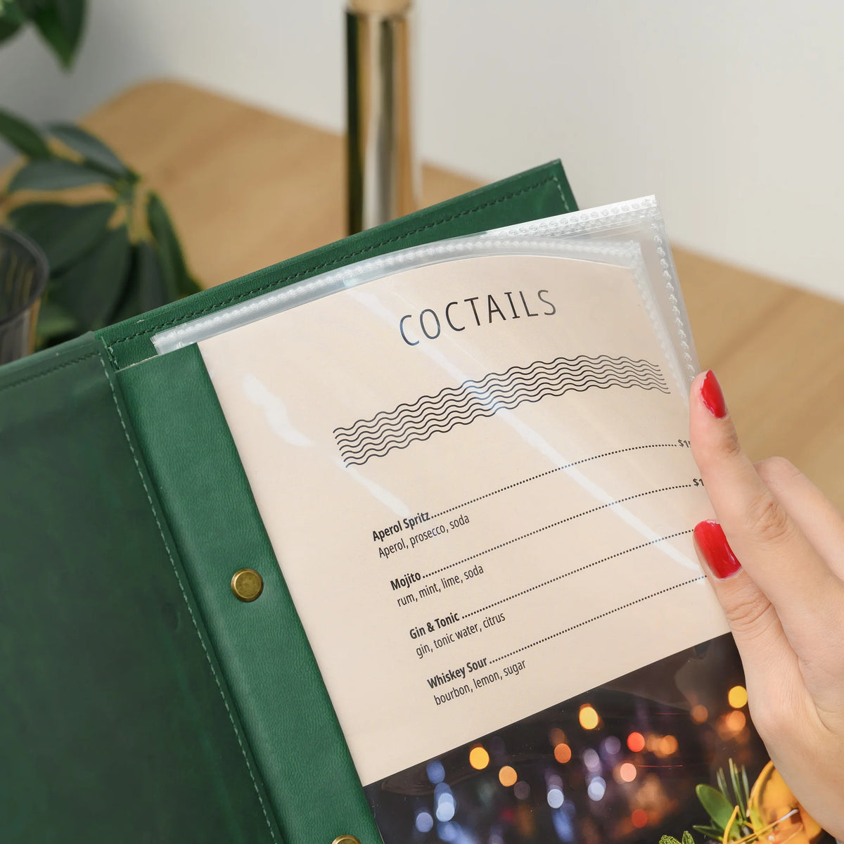Hand lifting the corner of a clear plastic sleeve in a green faux leather menu cover to show the printed menu page