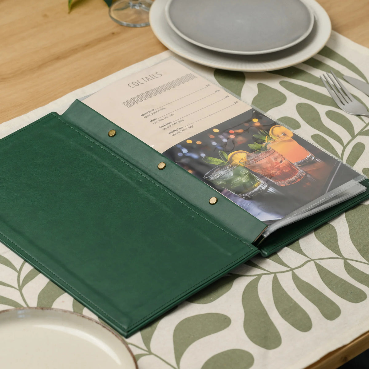 Open green faux leather menu cover lying on a set restaurant table with plates, showing the menu in a clear sleeve