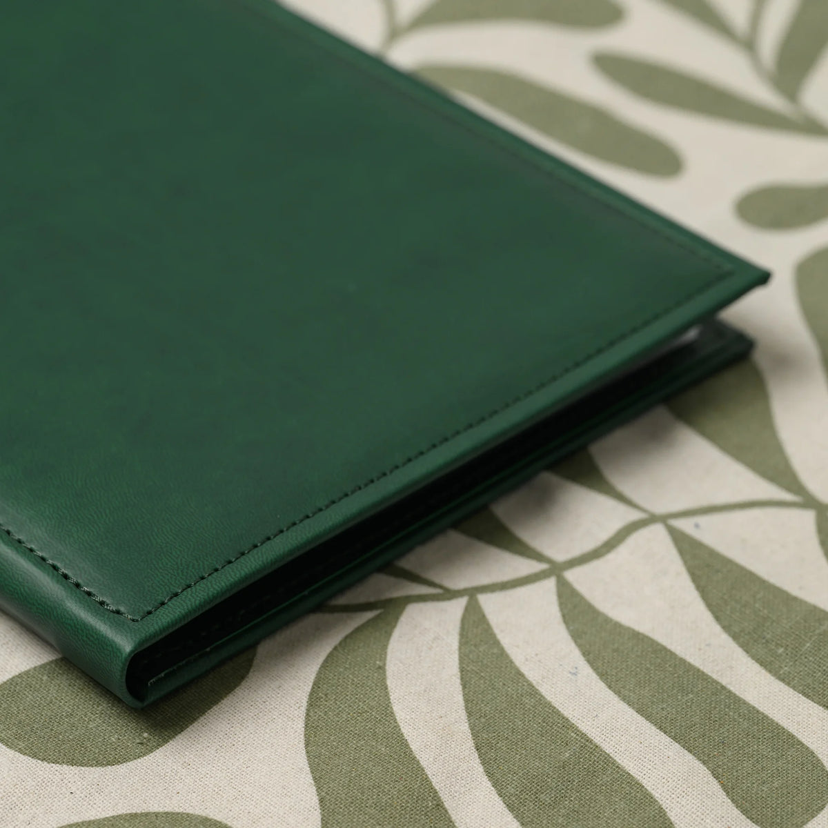 Close-up of the stitched edge and rounded corner of a closed green faux leather menu cover on a patterned runner