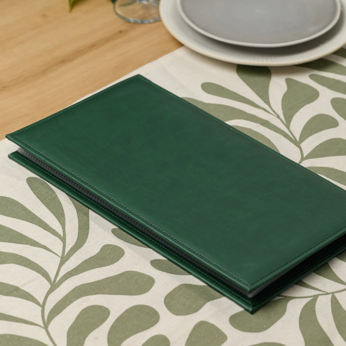 Closed green faux leather menu cover on a table runner, with the edges of the plastic sleeves visible inside it