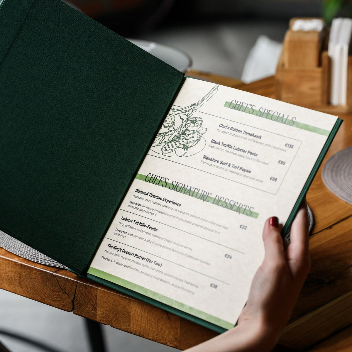 Hand holds an open dark green textile menu folder with a cream page of printed menu text on wooden table