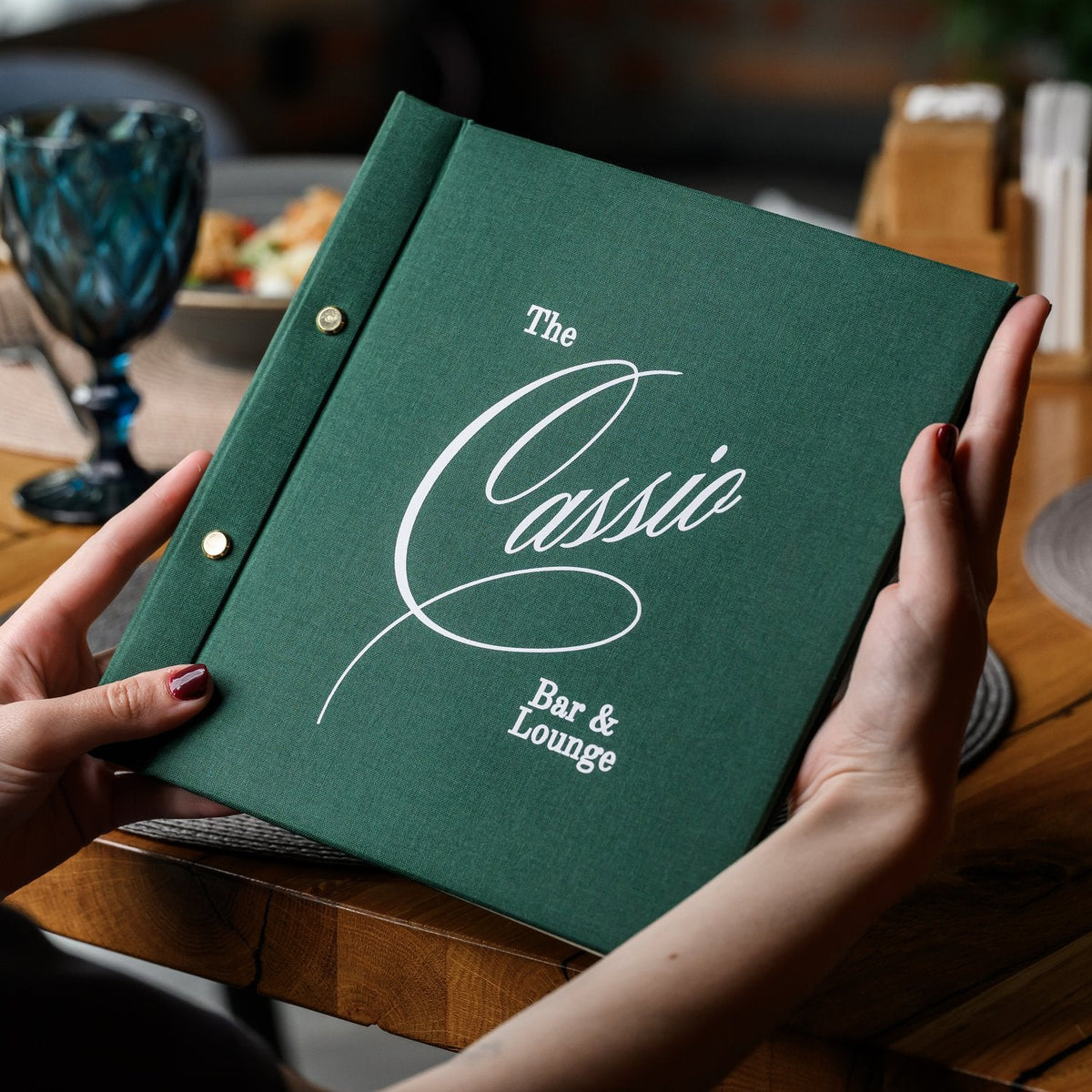 Hand holds a closed dark green textile menu folder with a script logo beside a blurred salad plate and wine