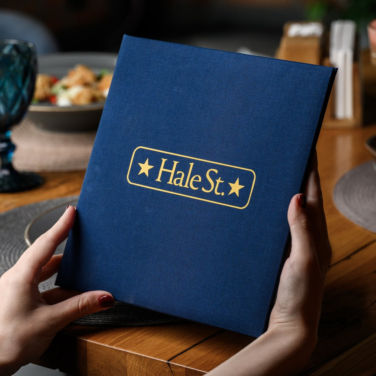 Two hands hold a closed navy textile menu folder with a gold star-badge logo above a set restaurant table