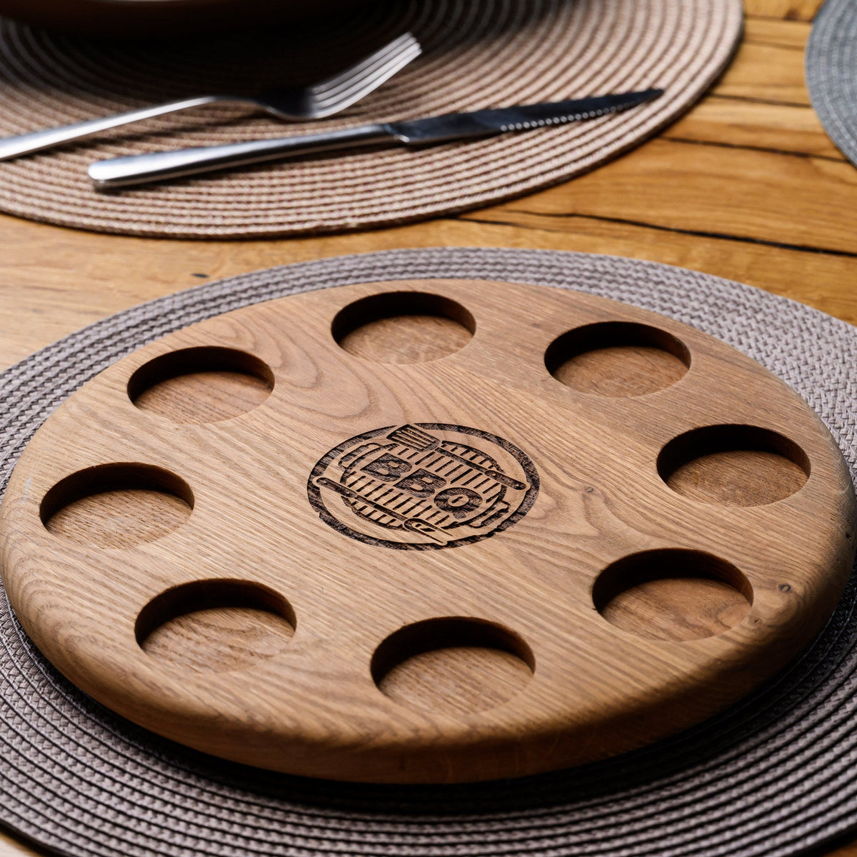 Inko oak BBQ serving board on woven grey placemat, angled view showing depth of shot glass recesses and burnt engraving