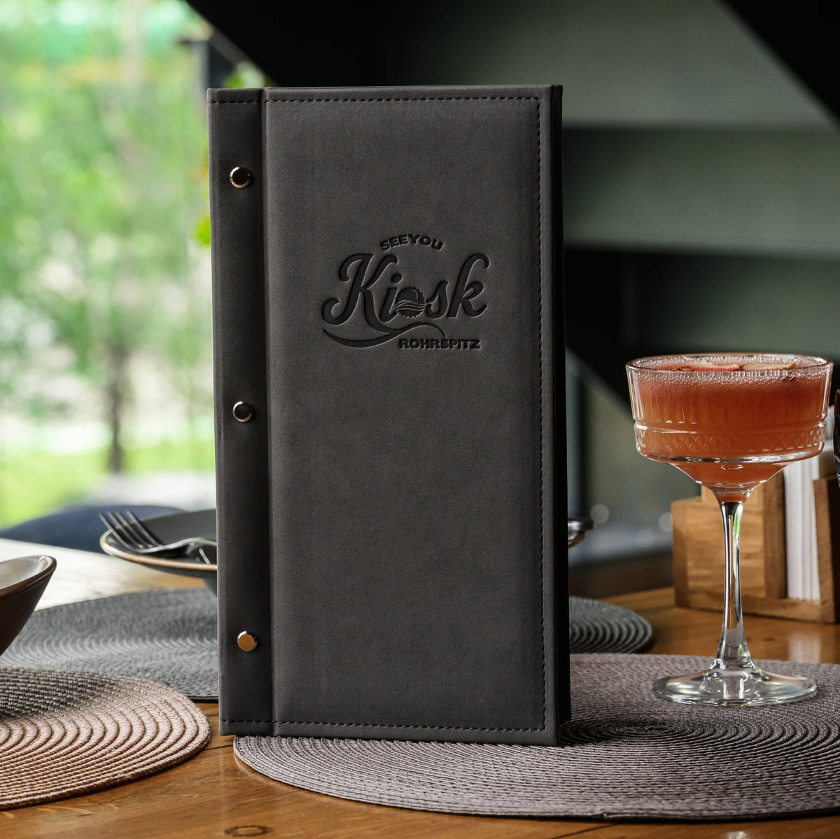 Closed nubuk menu cover standing upright with debossed Kiosk Rohrspitz logo beside cocktails on table