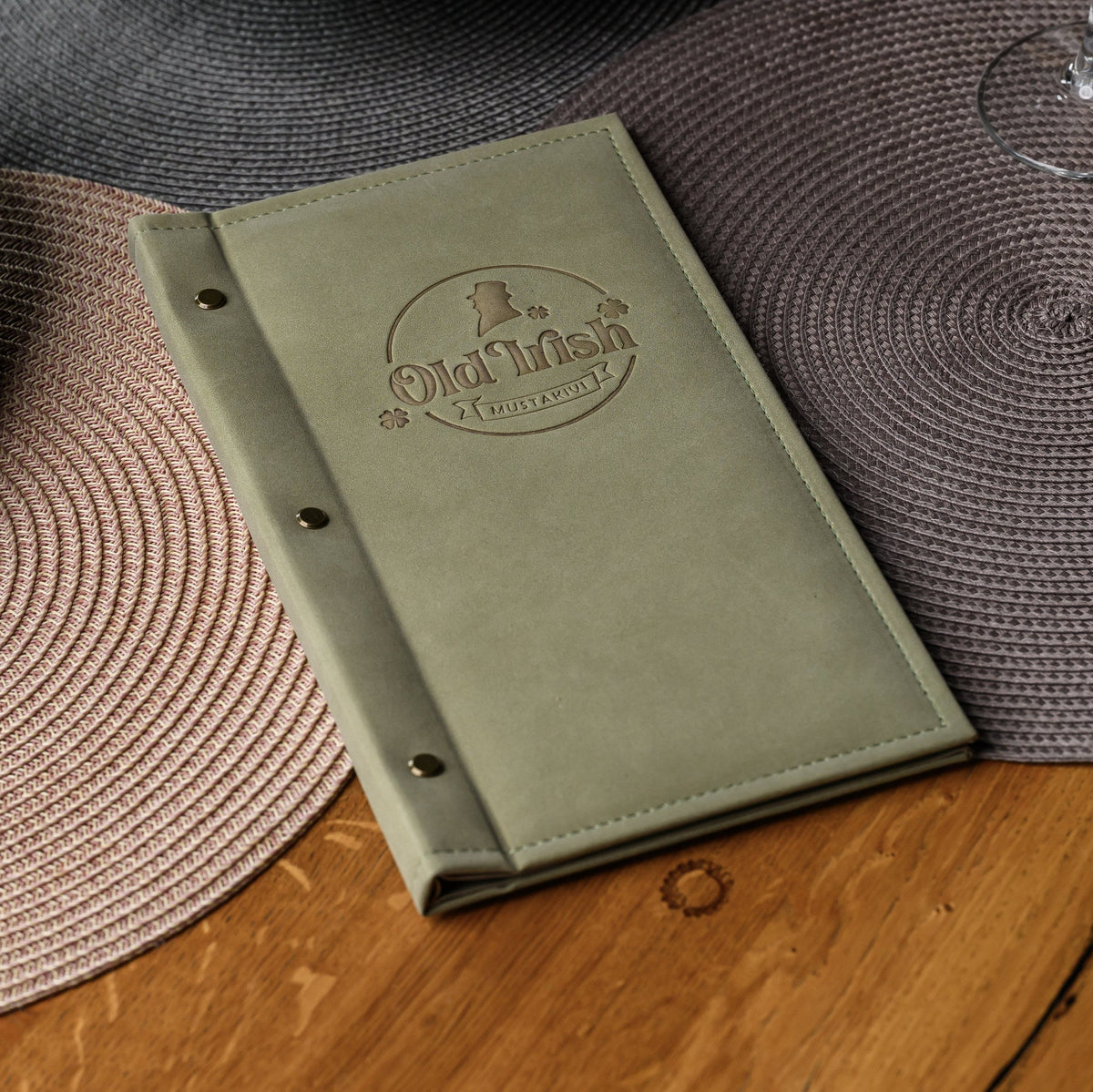 Closed olive nubuk menu cover on woven placemat with debossed Old Irish Mustarivi brand mark, wide view