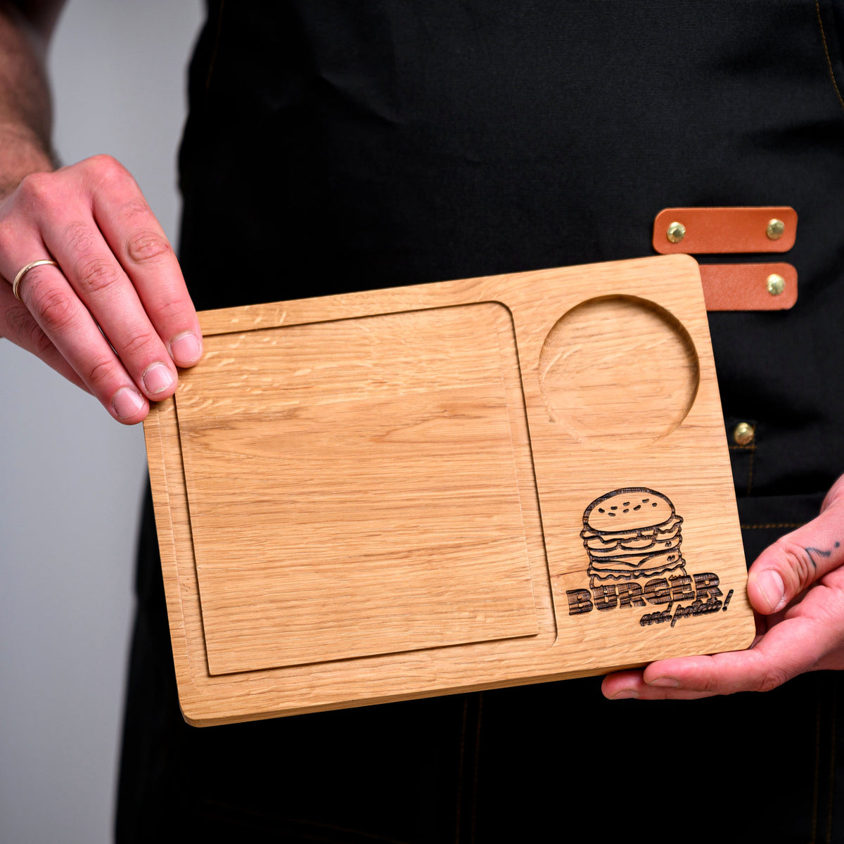 Oak two-section serving board with burger engraving held in dark apron, showing rectangular tray and sauce recess