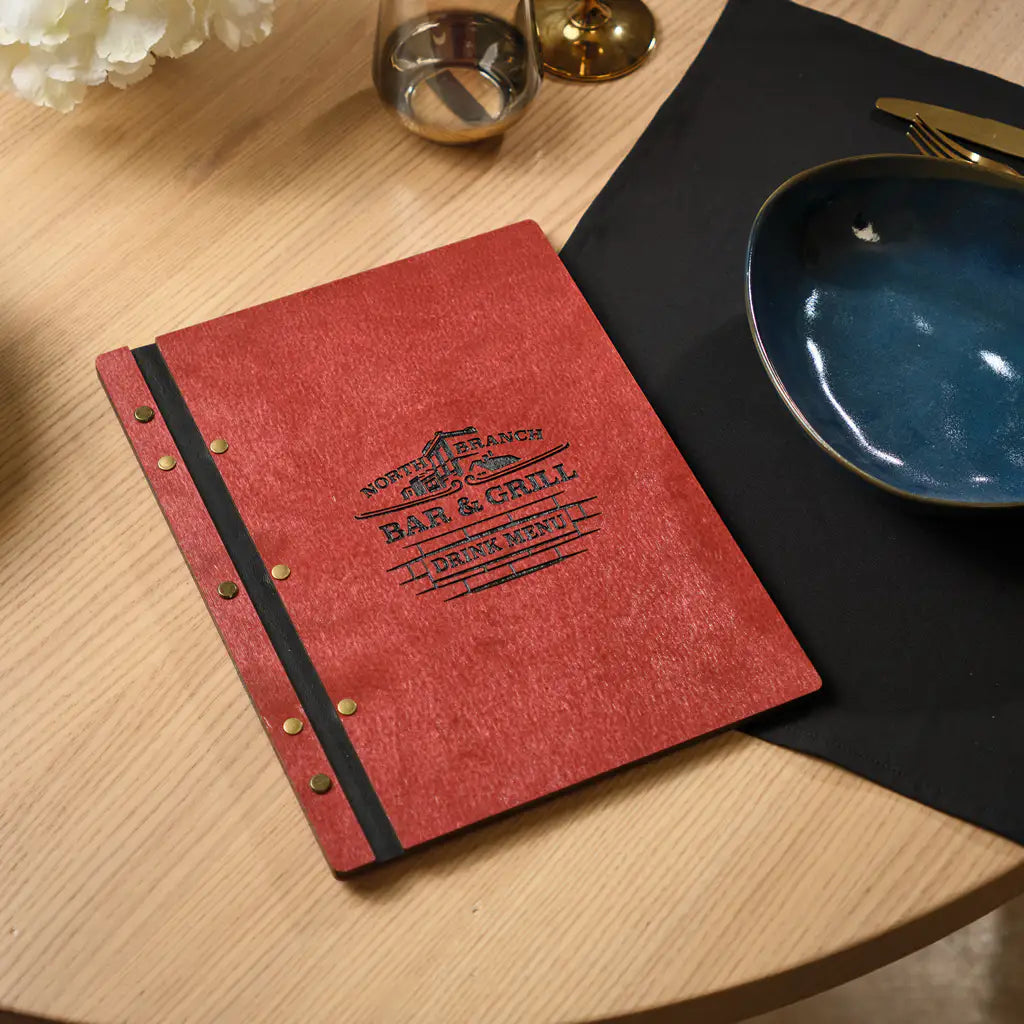 Closed chili red plywood menu folder standing on a wooden restaurant table. The cover shows a rich deep red painted plywood surface with a vertical black elastic band and brass snap button closure. An embossed brand logo is visible at the bottom of the cover. A dark blue ceramic bowl and golden cutlery are in the background. Stylish and durable plywood menu cover for upscale cafes, bistros and restaurants.