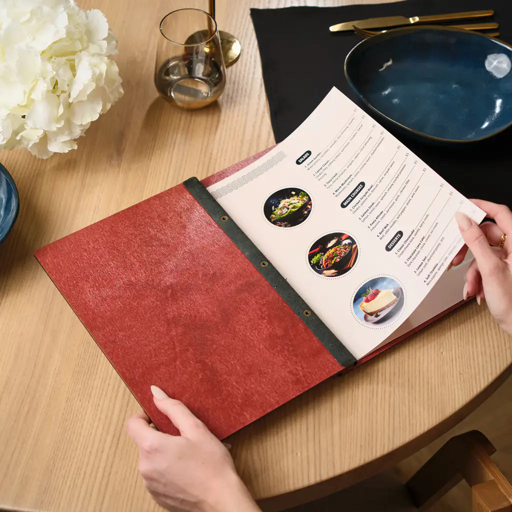 Chili red plywood restaurant menu folder opened by hands on a round wooden table. The cover features a black elastic band with brass metal snap buttons on the spine. A printed menu insert with food photos is visible inside. White flowers and a dark blue ceramic bowl are seen in the background. Bold colored plywood menu holder with durable snap button closure for restaurants, cafes and bars seeking a vibrant and modern table presentation.