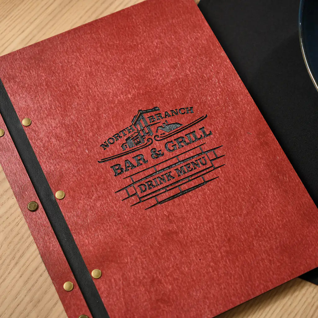 chili red plywood menu folder showing a custom engraved Bar and Grill logo on the inner right panel. A printed food menu insert is placed on the left side. The cover is flat on a wooden table with brass snap buttons and black elastic band visible along the spine. Personalized plywood menu holder with custom logo engraving for branded restaurant, bar and grill table settings.