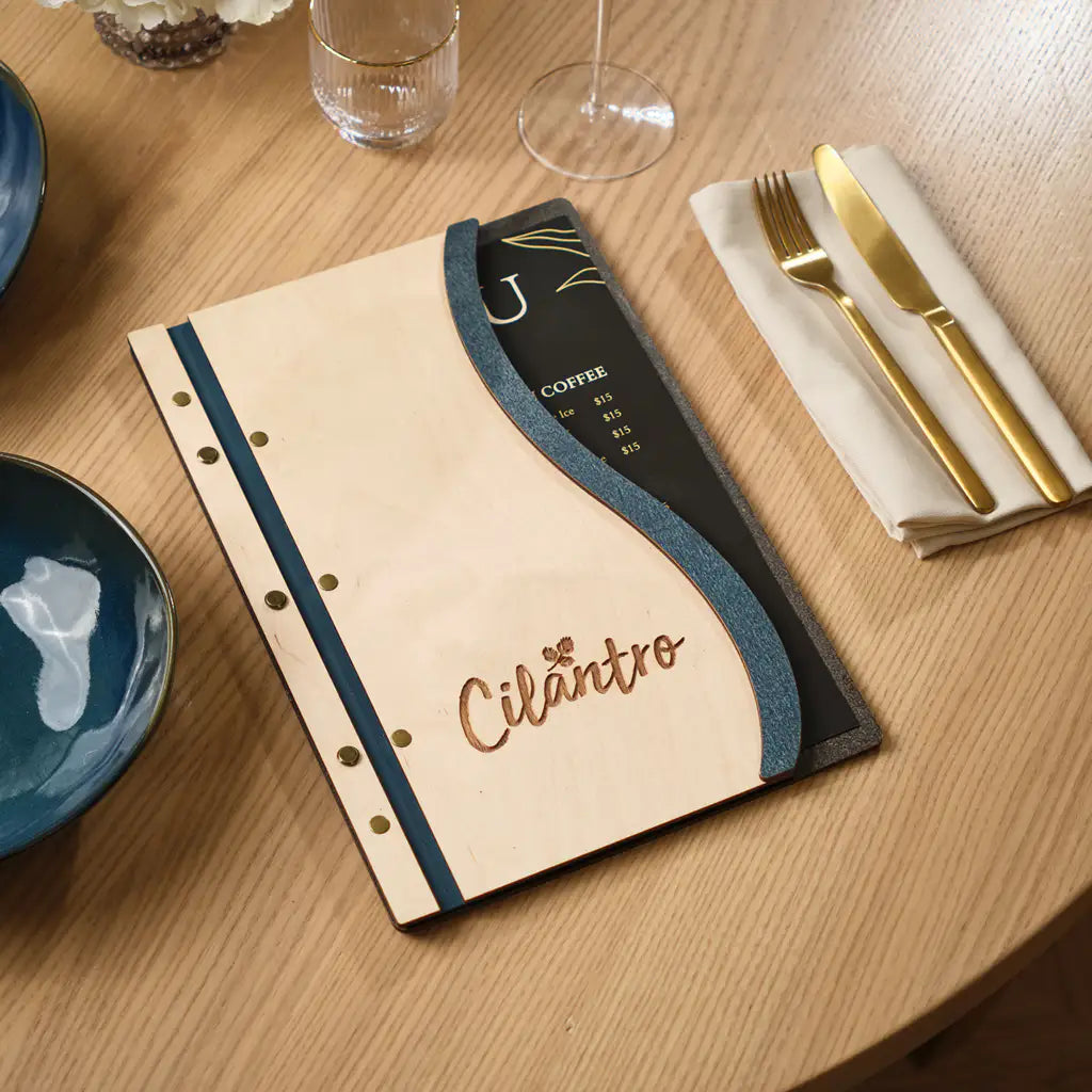 Slim plywood menu folder with denim wave insert and engraved Cilantro logo on wooden restaurant table
