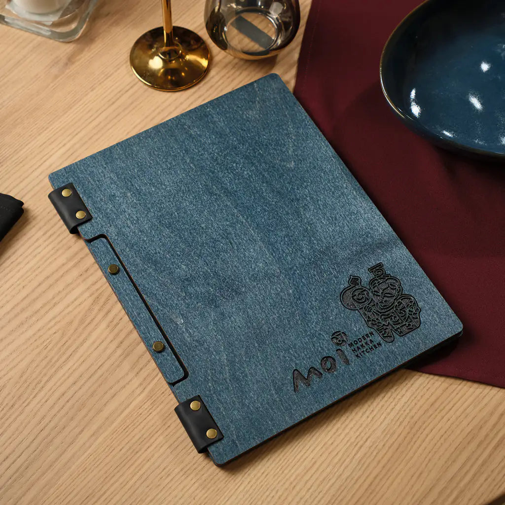 Closed denim blue plywood square menu folder with engraved Moi logo, gold candlesticks in background
