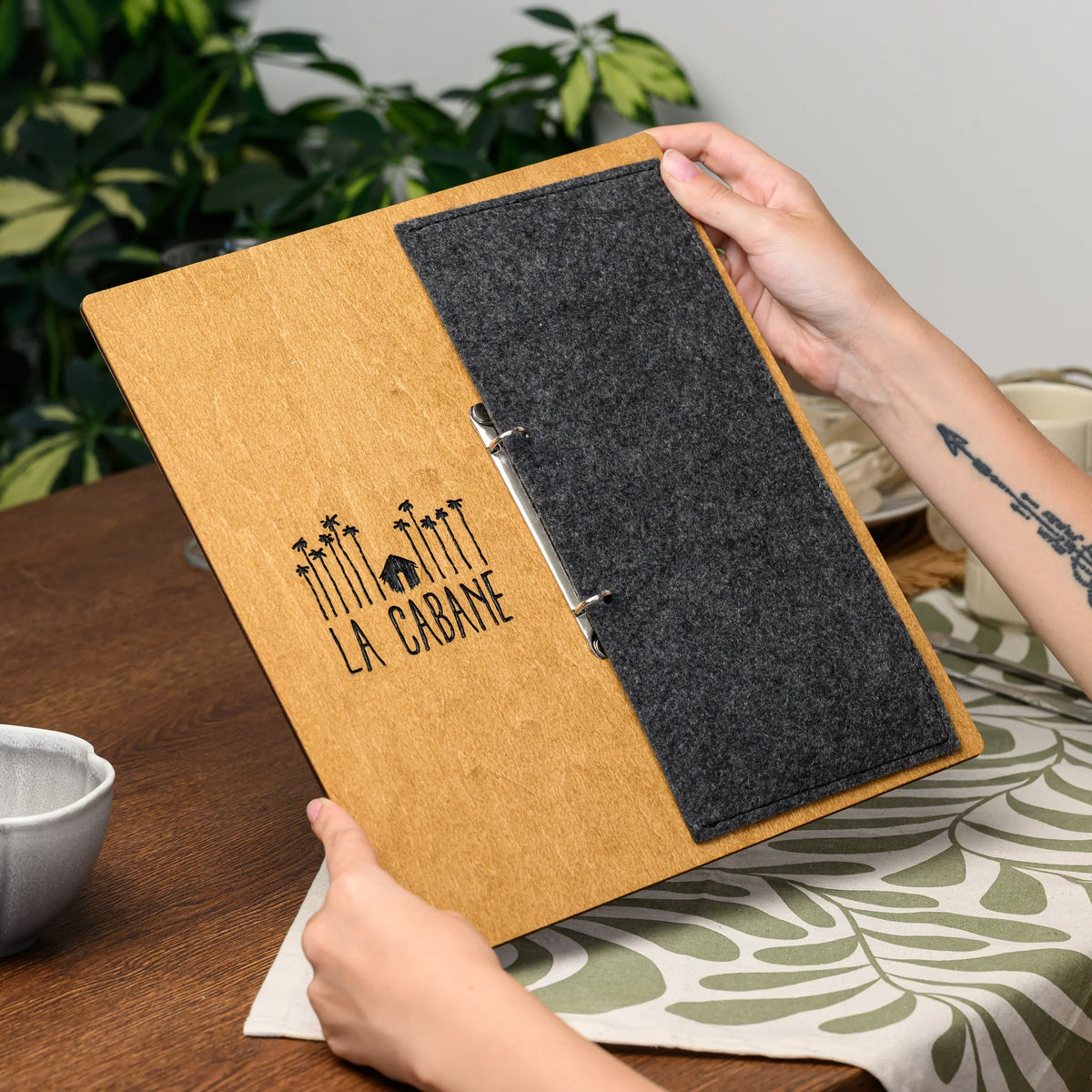 Hands holding a closed menu board, showing the engraved plywood front and felt cover panel near a plant 